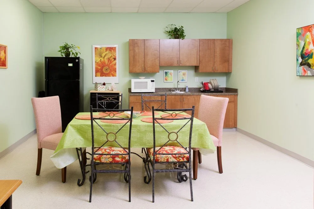 A kitchen and dining area with a table covered with a green tablecloth, four chairs, a black refrigerator, a microwave, wooden cabinets, and colorful wall art.
