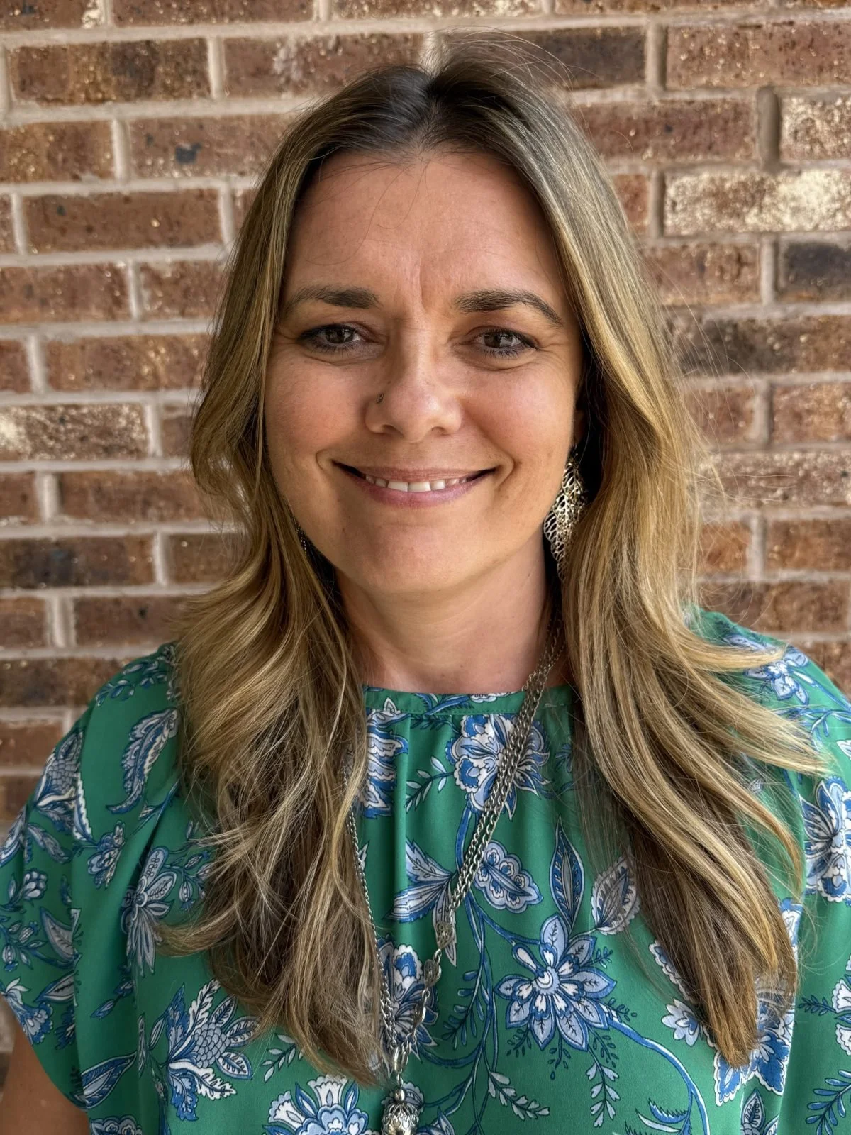 A woman with long wavy blonde hair smiling in front of a brick wall, wearing a green floral dress, earrings, and a necklace.