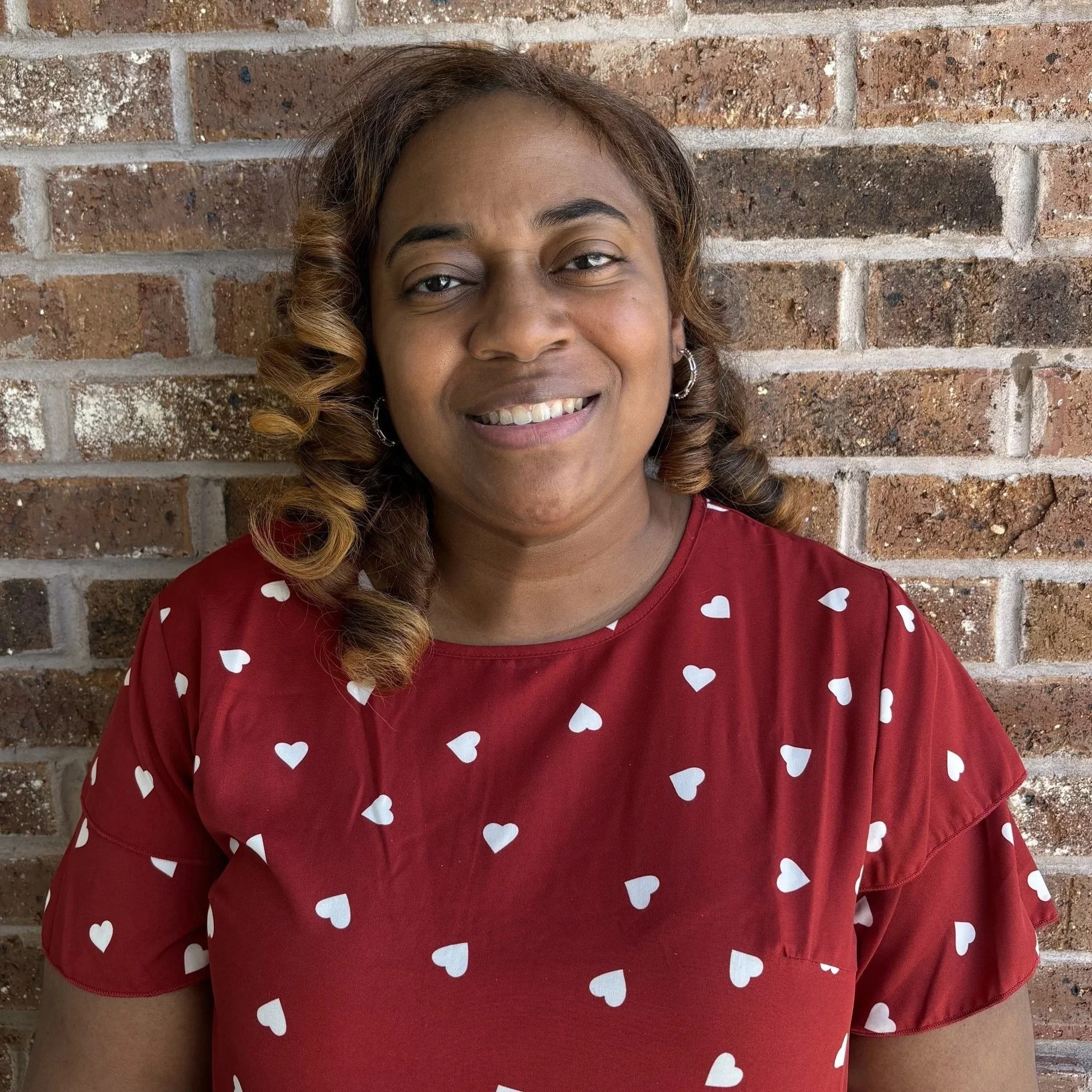 A woman smiling in front of a brick wall, wearing a leopard print top and having long blonde braided hair.