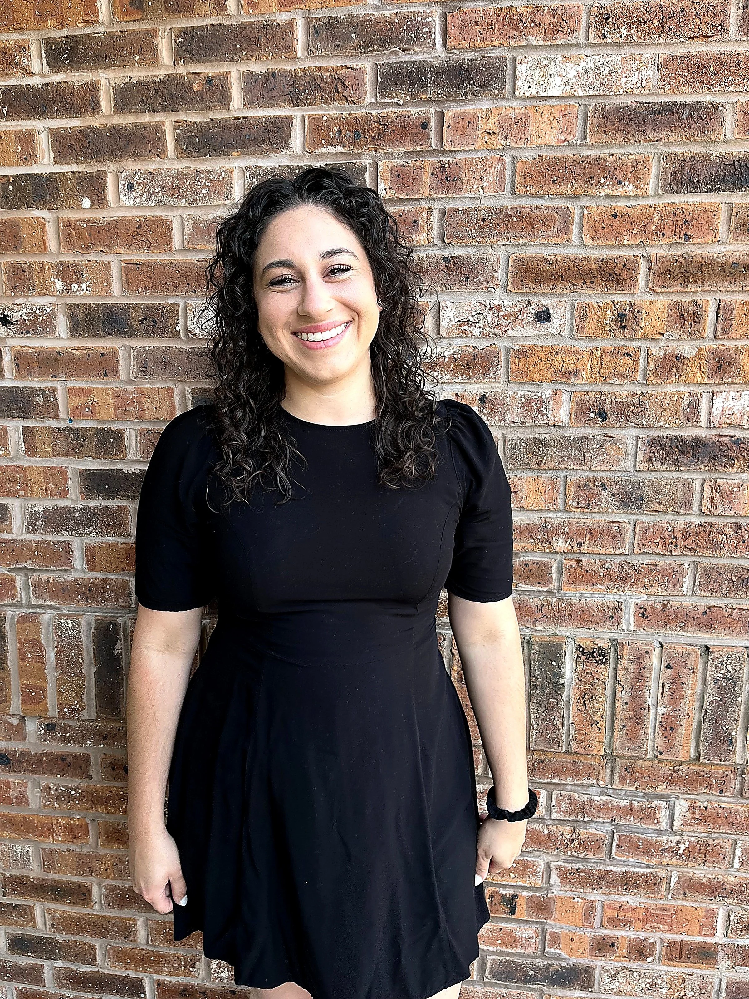 A young woman with curly dark hair, smiling, wearing a black dress with puffed sleeves, standing in front of a brick wall.
