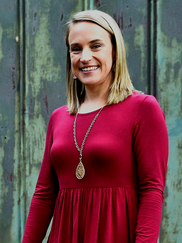 A woman with shoulder-length blonde hair, smiling, wearing a red dress with long sleeves, accessorized with a long gold chain necklace with a pendant, standing in front of a weathered green and gray metal wall.