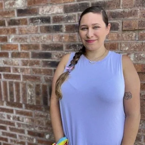 Young woman with long hair in a braid, wearing a sleeveless light blue top, standing in front of a brick wall, smiling with a patch tattoo on her upper arm.