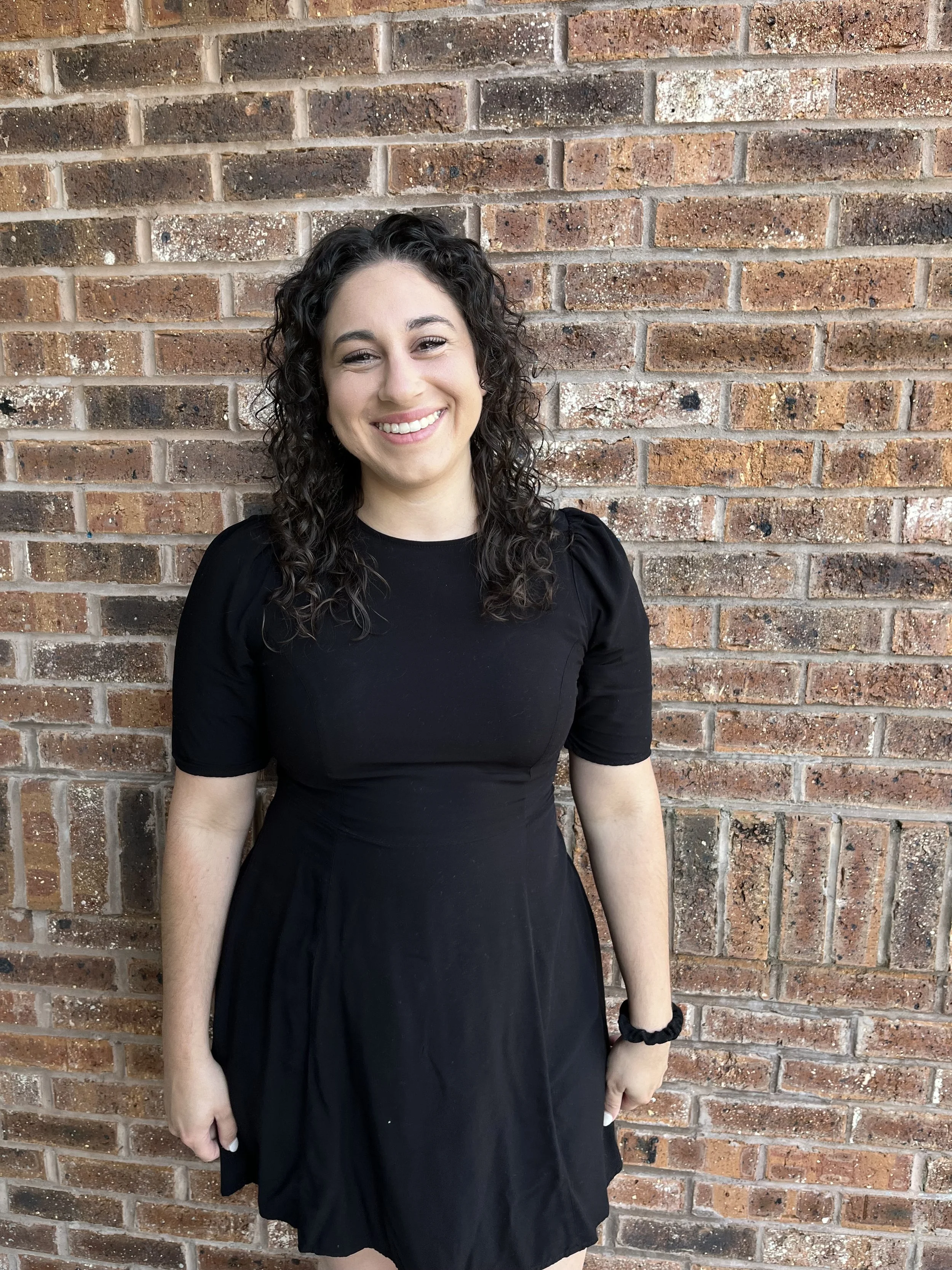 A young woman with curly dark hair, wearing a black dress and a black hair tie, standing in front of a brick wall and smiling at the camera.