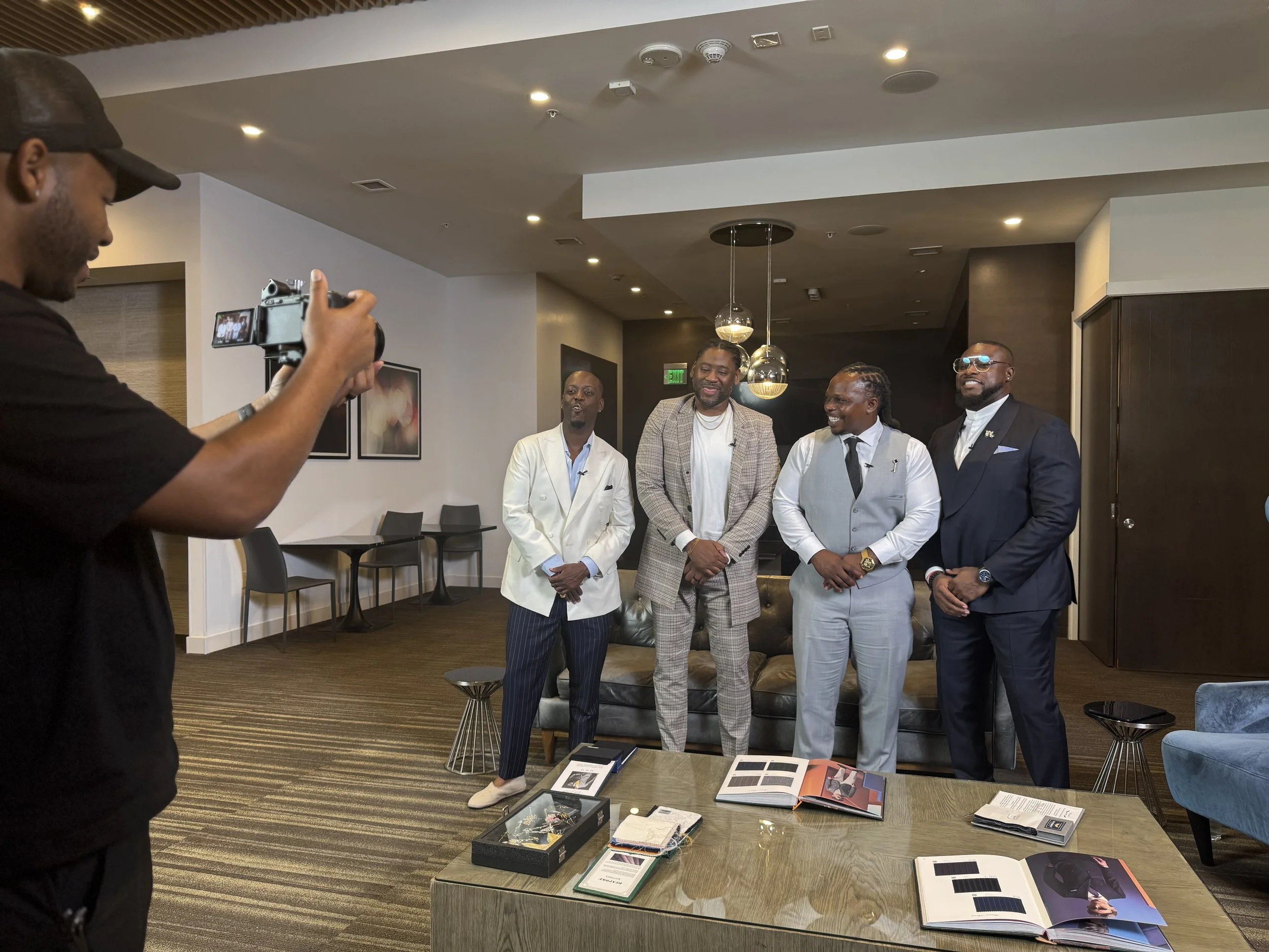 Four men in formal and semi-formal attire posing for a photo in a modern, well-lit room with a man taking their picture.