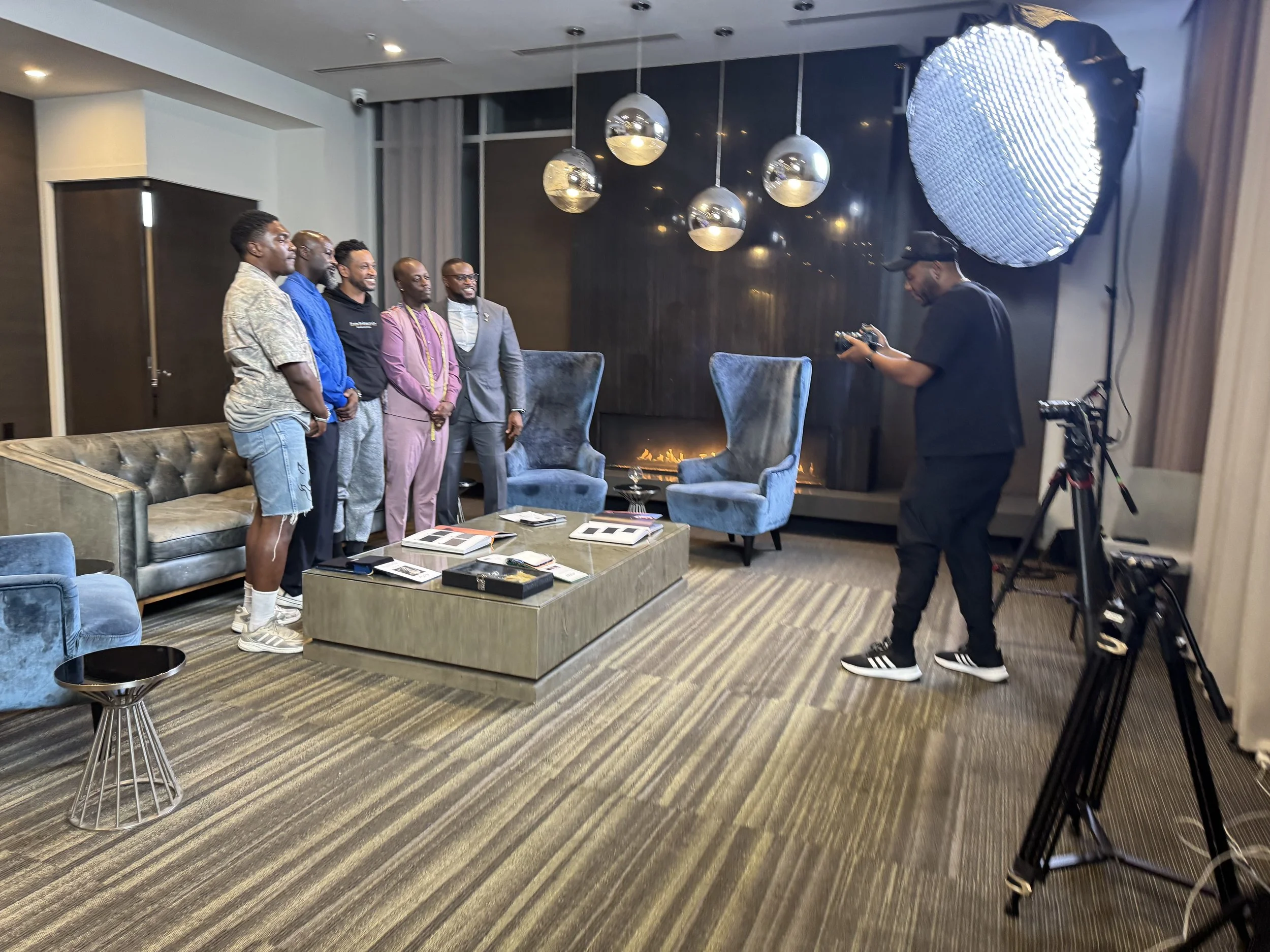 Six men standing together in a well-lit lounge area with modern decor, a large mirror, and picture books on the table, while a photographer takes their picture. The men are dressed in a mix of casual and formal attire.