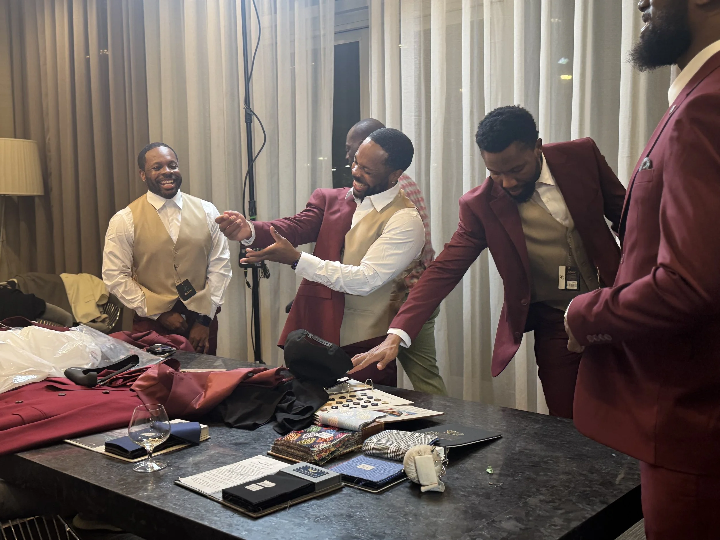 Group of men in formal attire smiling and engaging in conversation in a hotel room, with a table of clothing and accessories.