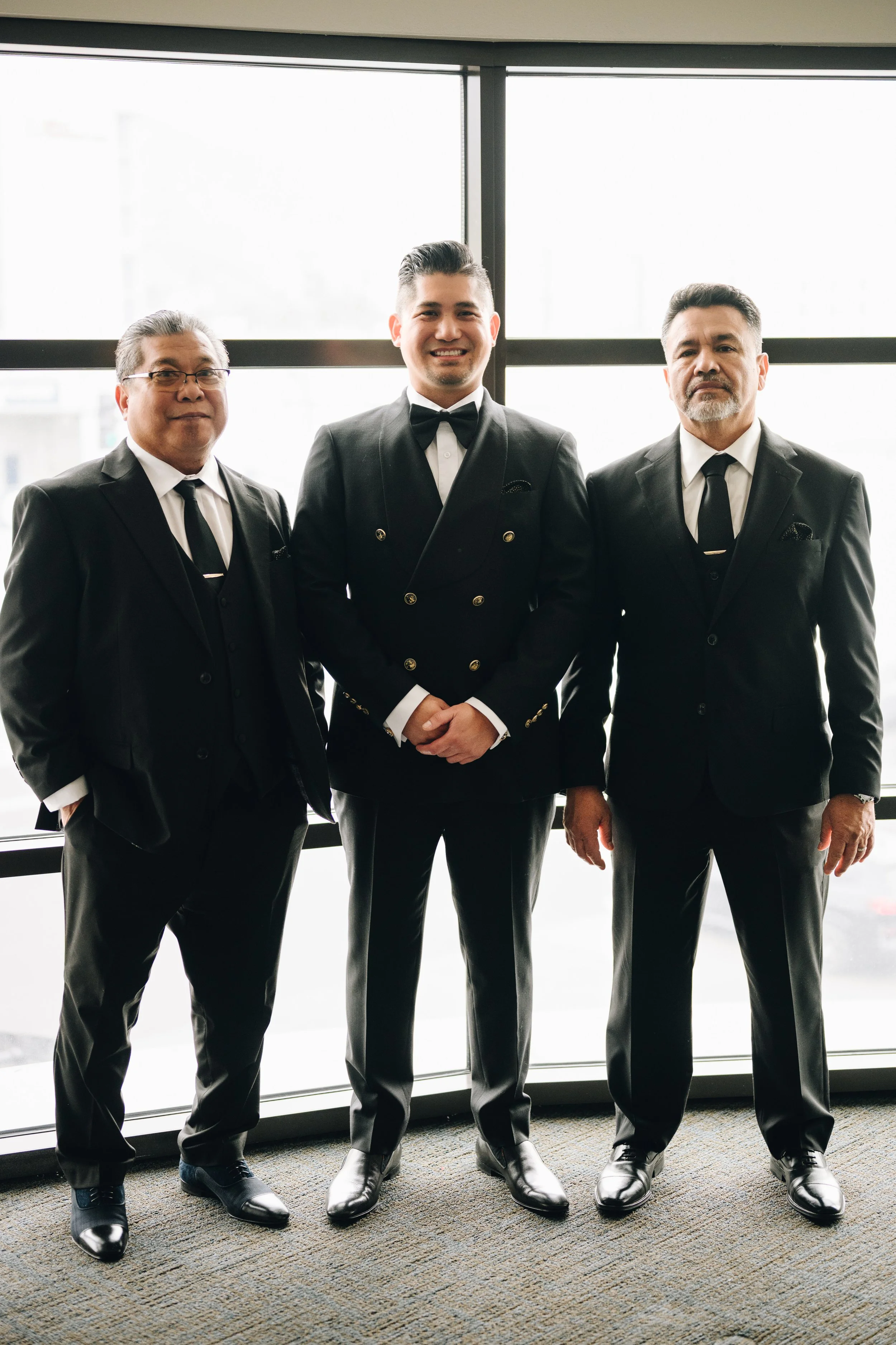 Three men in black tuxedos standing in front of a large window, smiling.