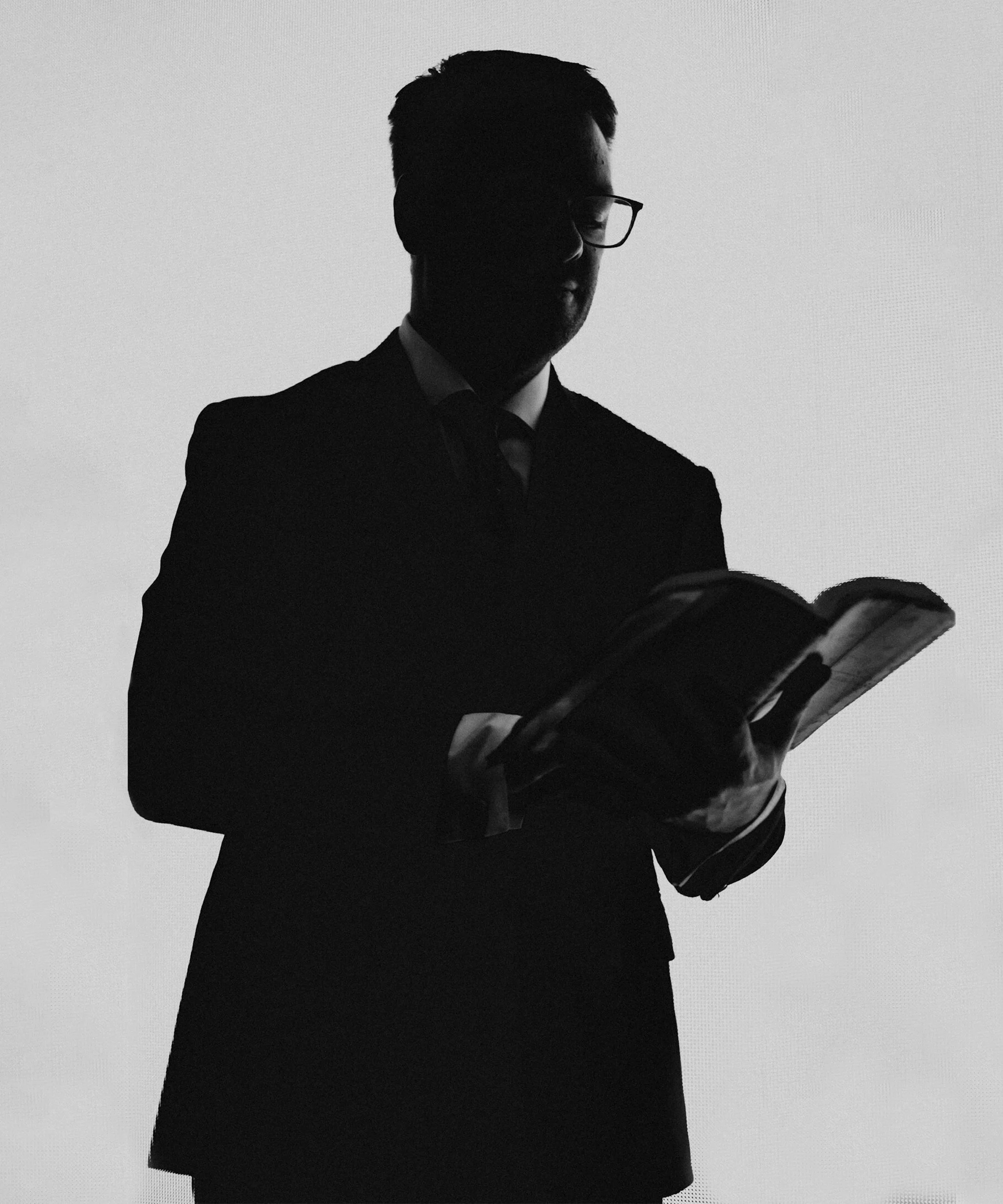 Silhouette of a man wearing glasses and a suit, holding an open book or magazine.