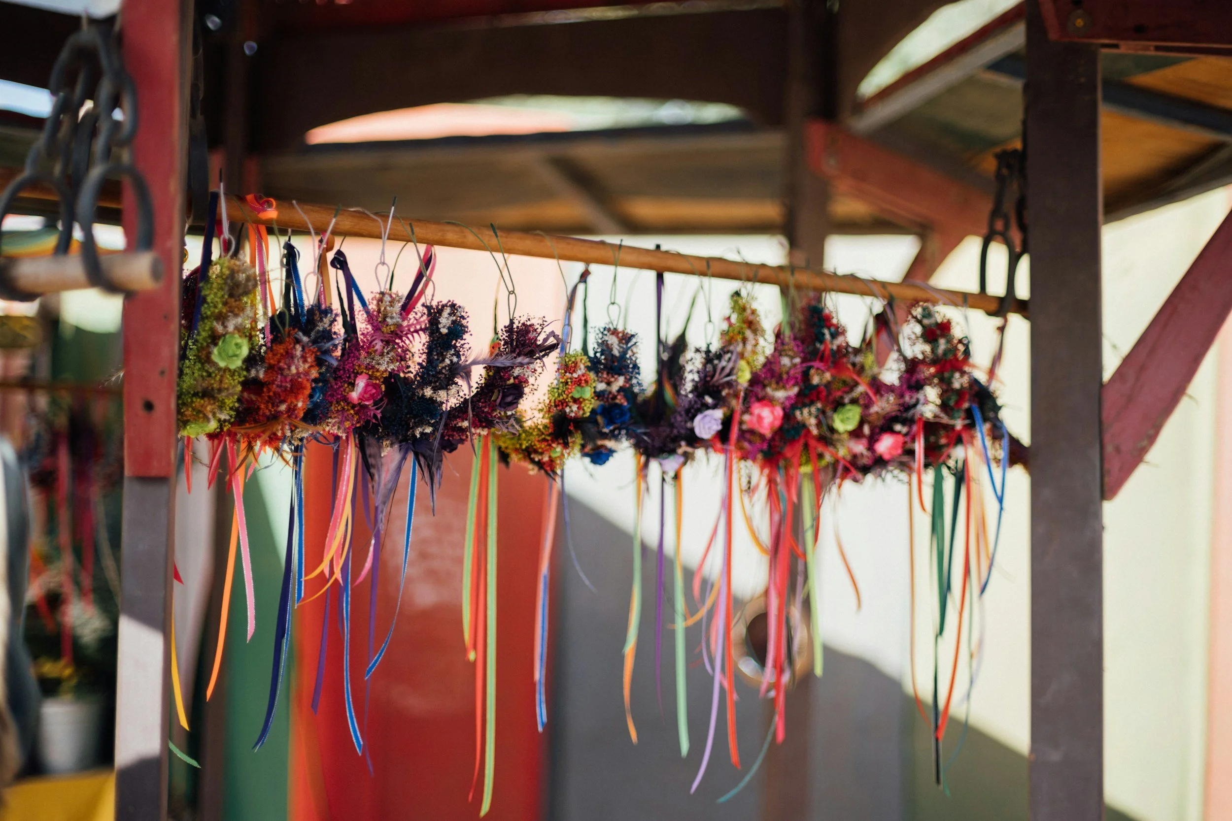 Colorful flower crowns hanging on a horizontal wooden rod at an outdoor market stall.