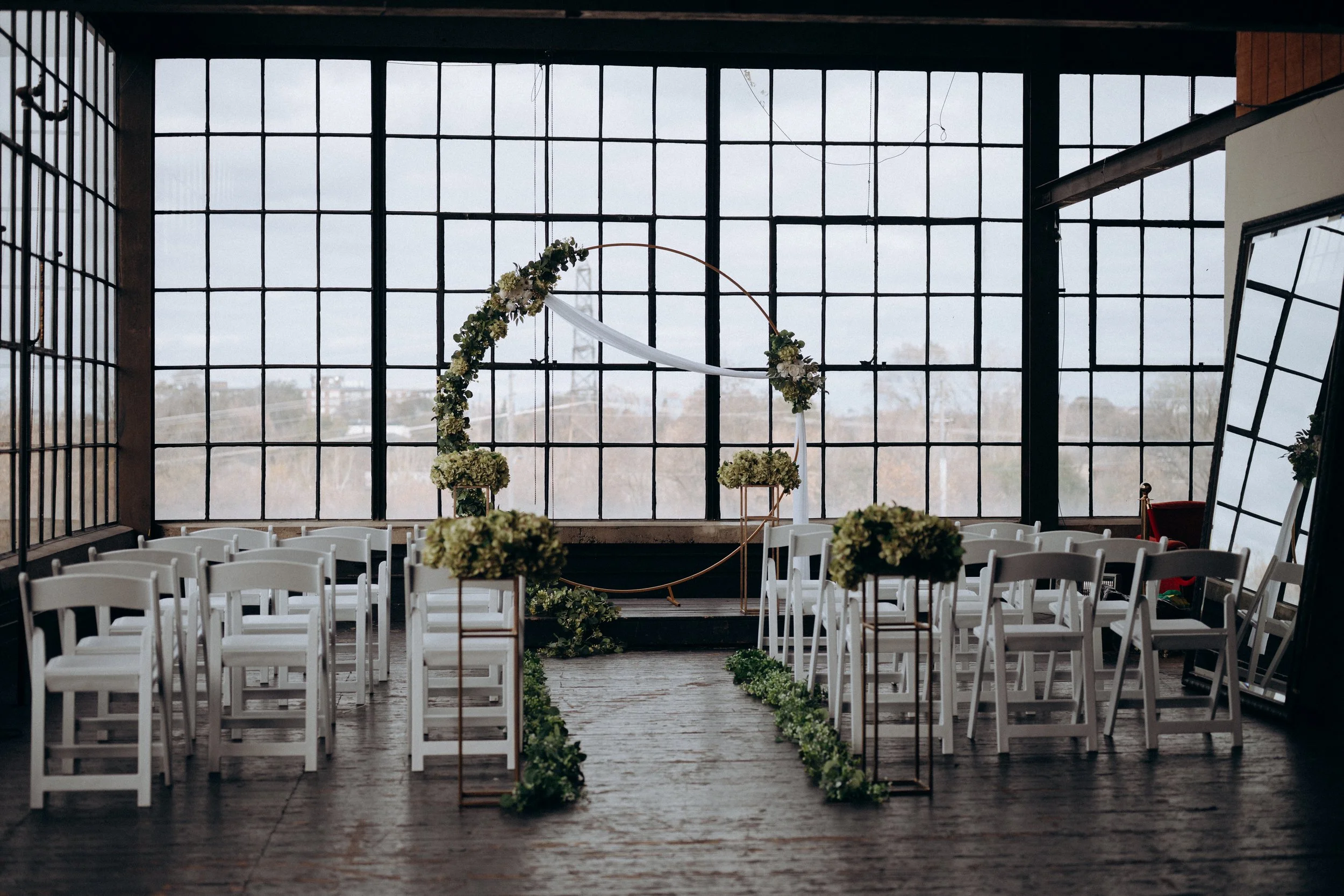 Indoor wedding ceremony setup with white chairs, floral arrangements, and a circular floral arch on a wooden floor, large windows in the background.