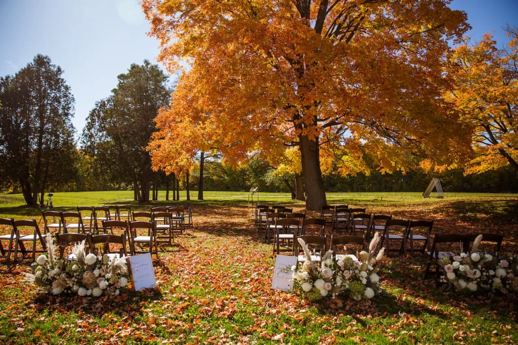 Outdoor wedding setup under autumn trees with chairs, floral arrangements, and signs.