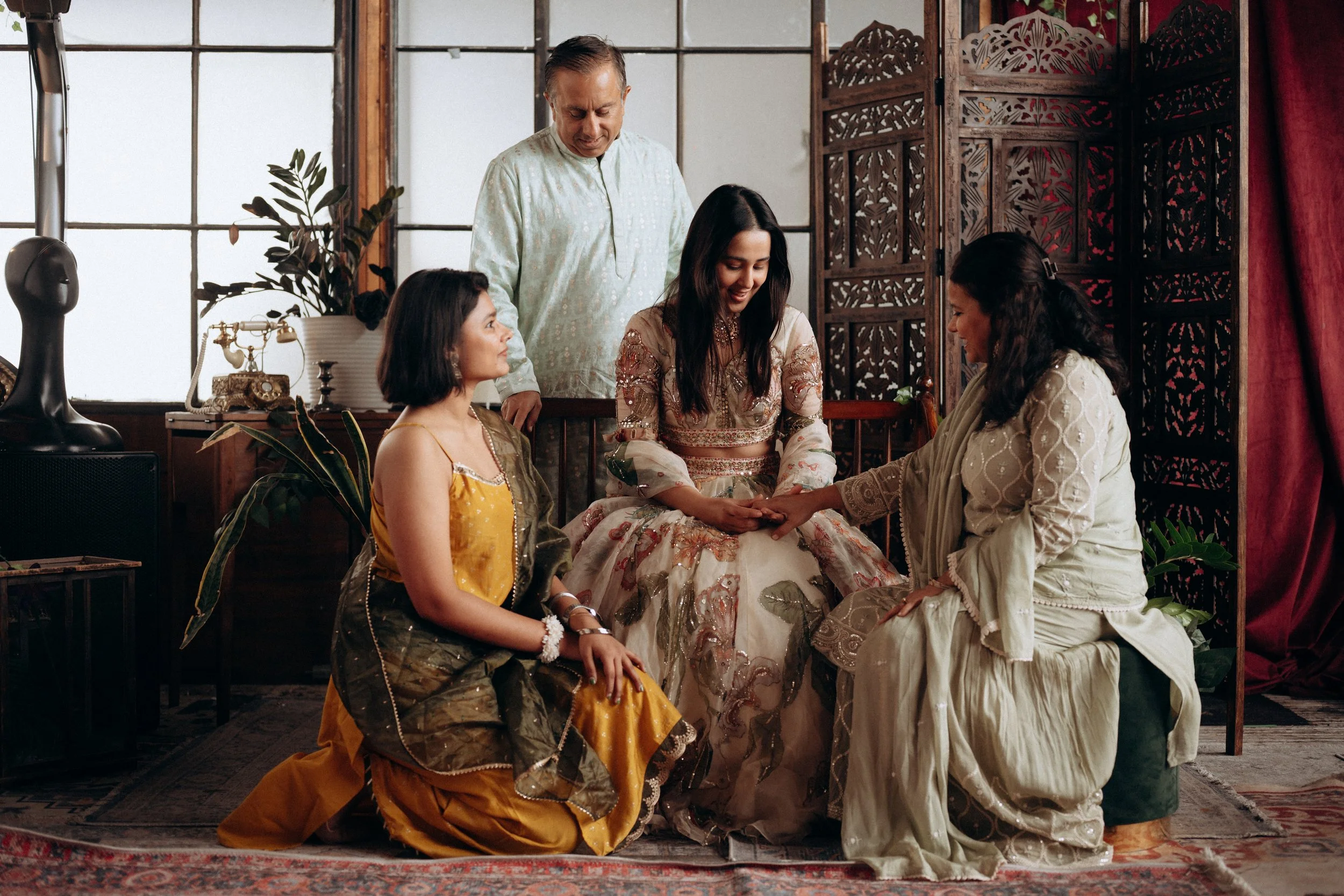 A woman in a floral traditional dress is seated, holding hands with another woman in a cream-colored dress, during a cultural ceremony with three other women and a man gathered around in a decorated room with wooden panels, plants, and large windows.