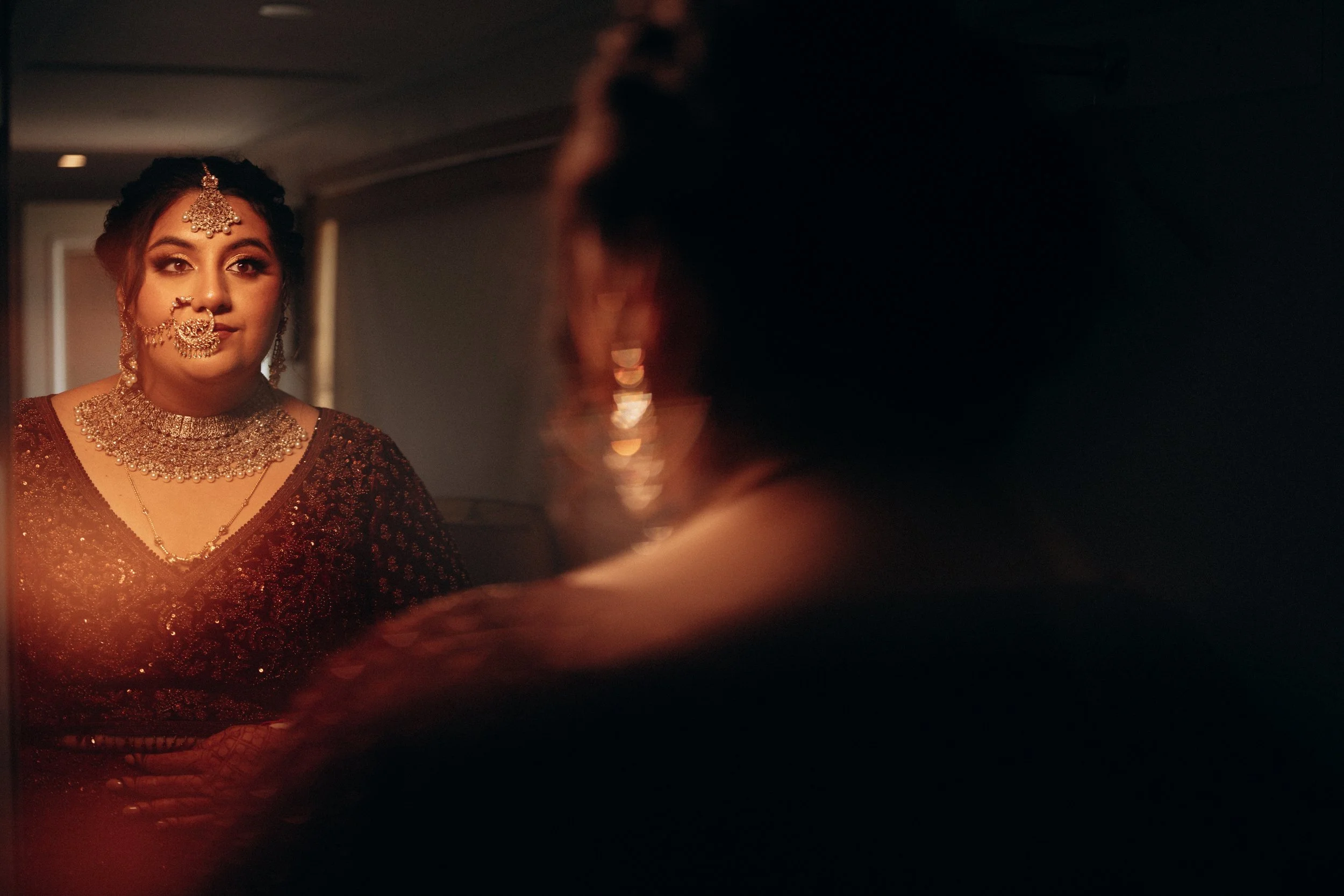A woman dressed in traditional Indian attire with jewelry, looking at herself in a mirror.