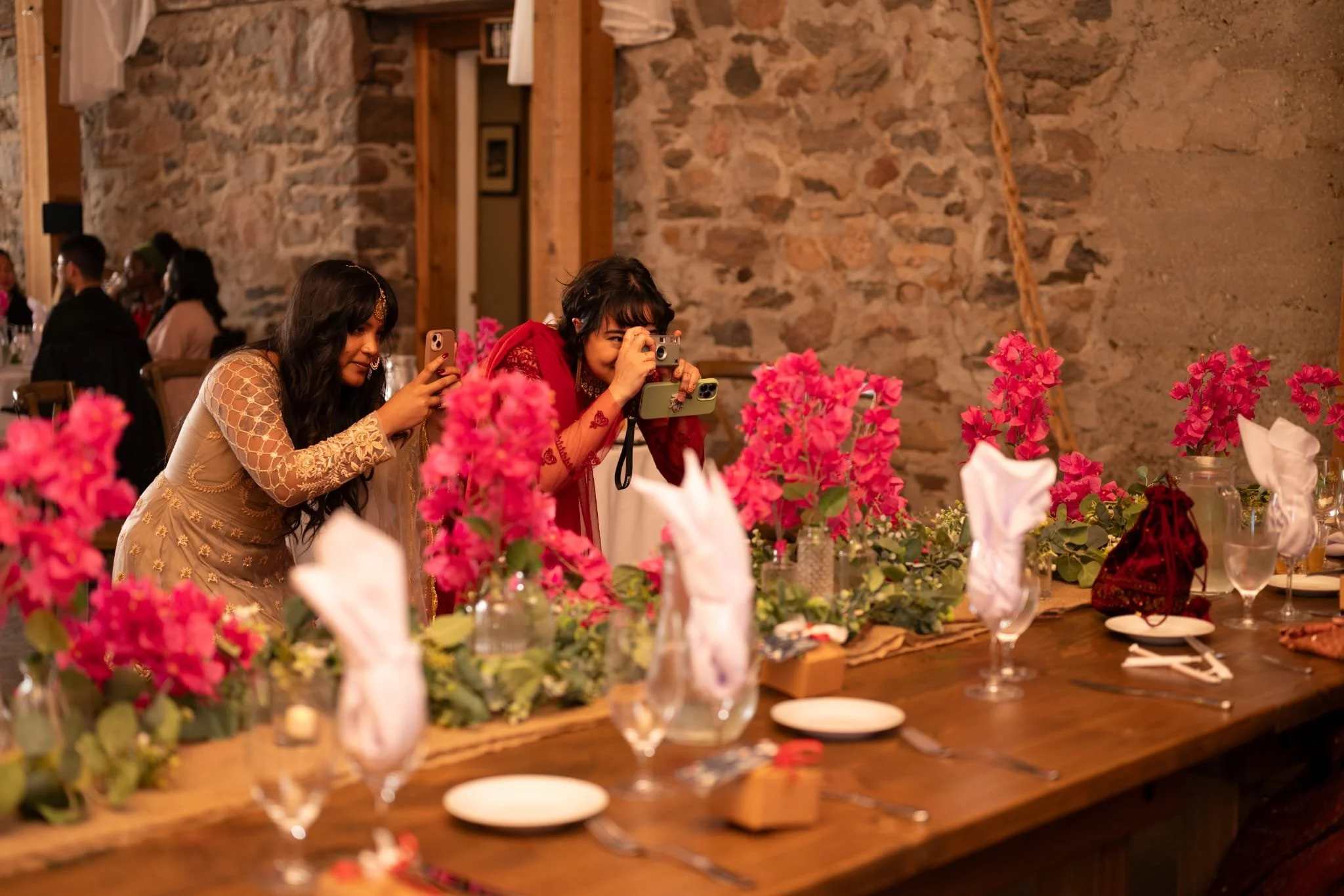 Two women in elegant dresses taking photos at a table decorated with pink flowers and white napkins in a rustic stone-walled setting.