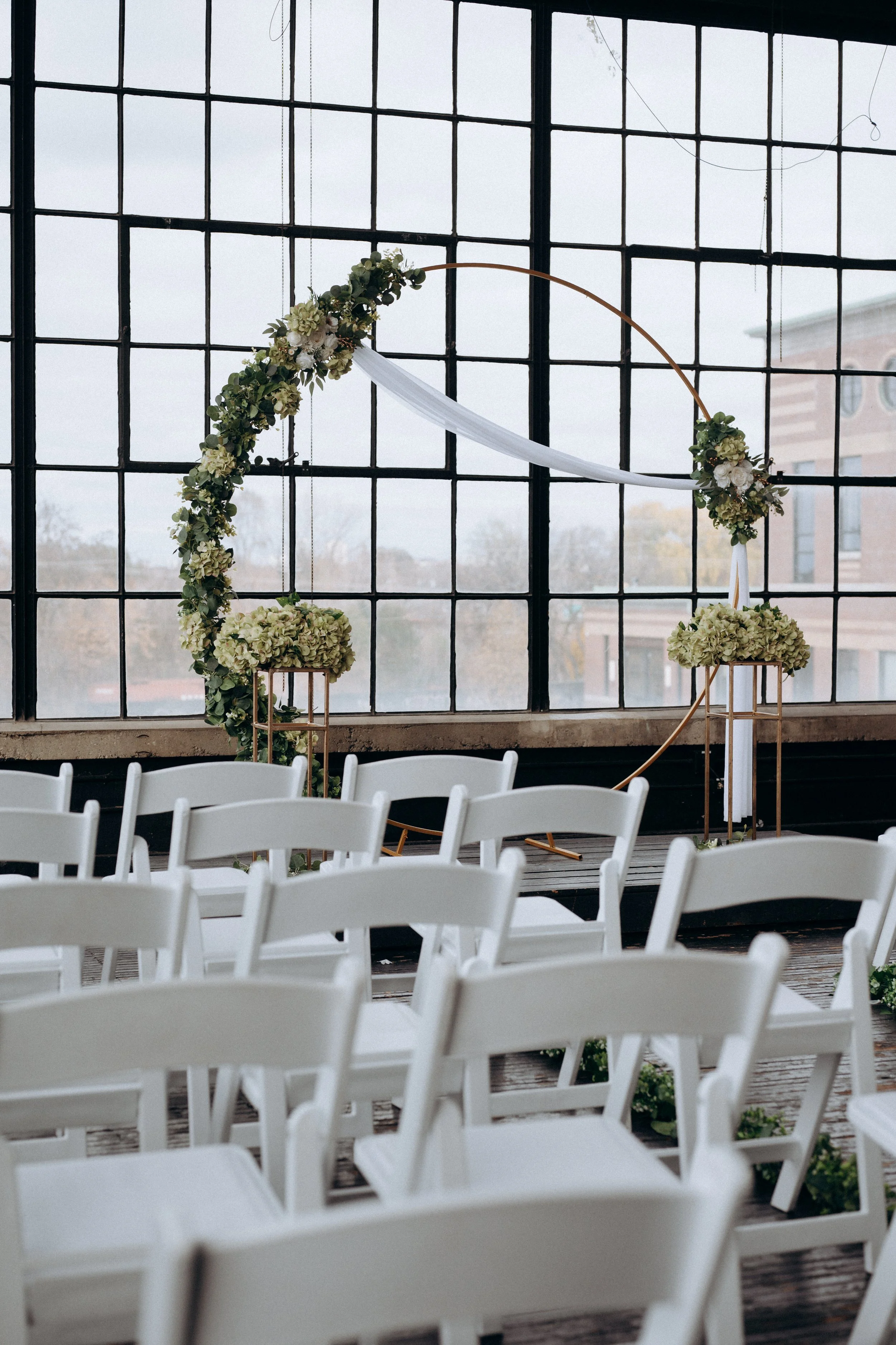 Wedding ceremony setup with white chairs facing a gold circular floral arch decorated with green and white flowers, in front of large industrial-style windows.