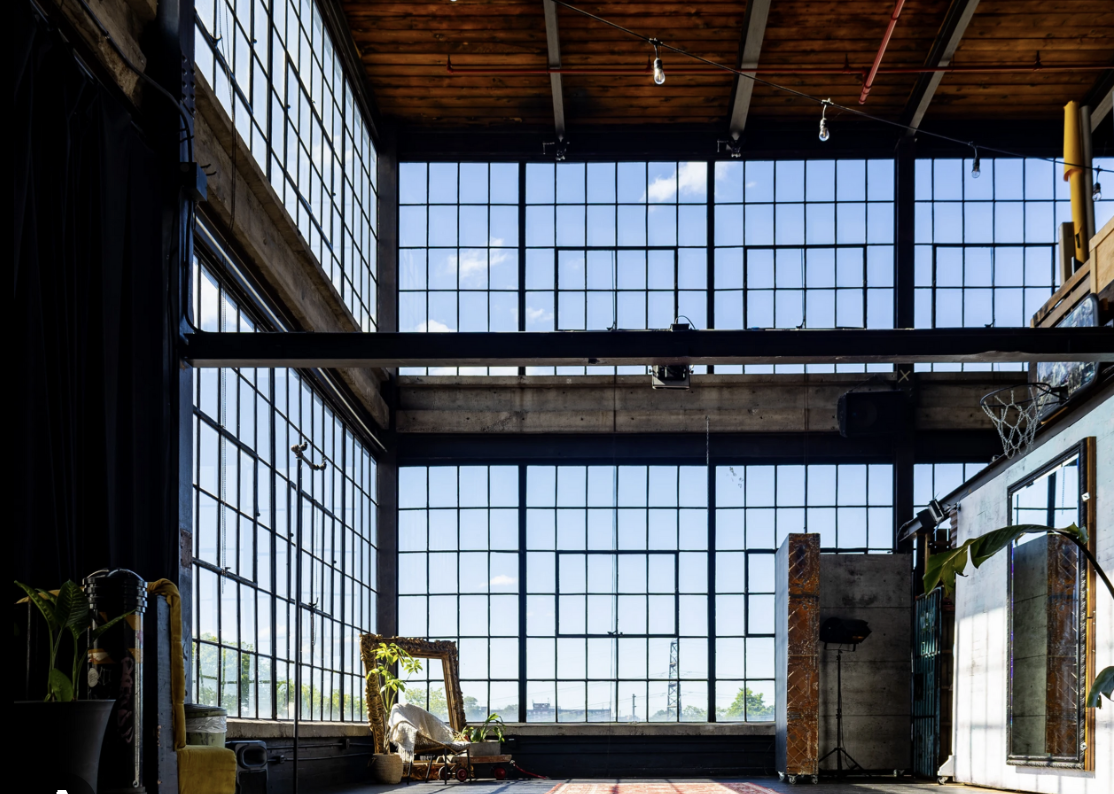 Interior of a spacious loft with large grid windows, exposed wooden ceiling, plants, vintage furniture, and decorative items.