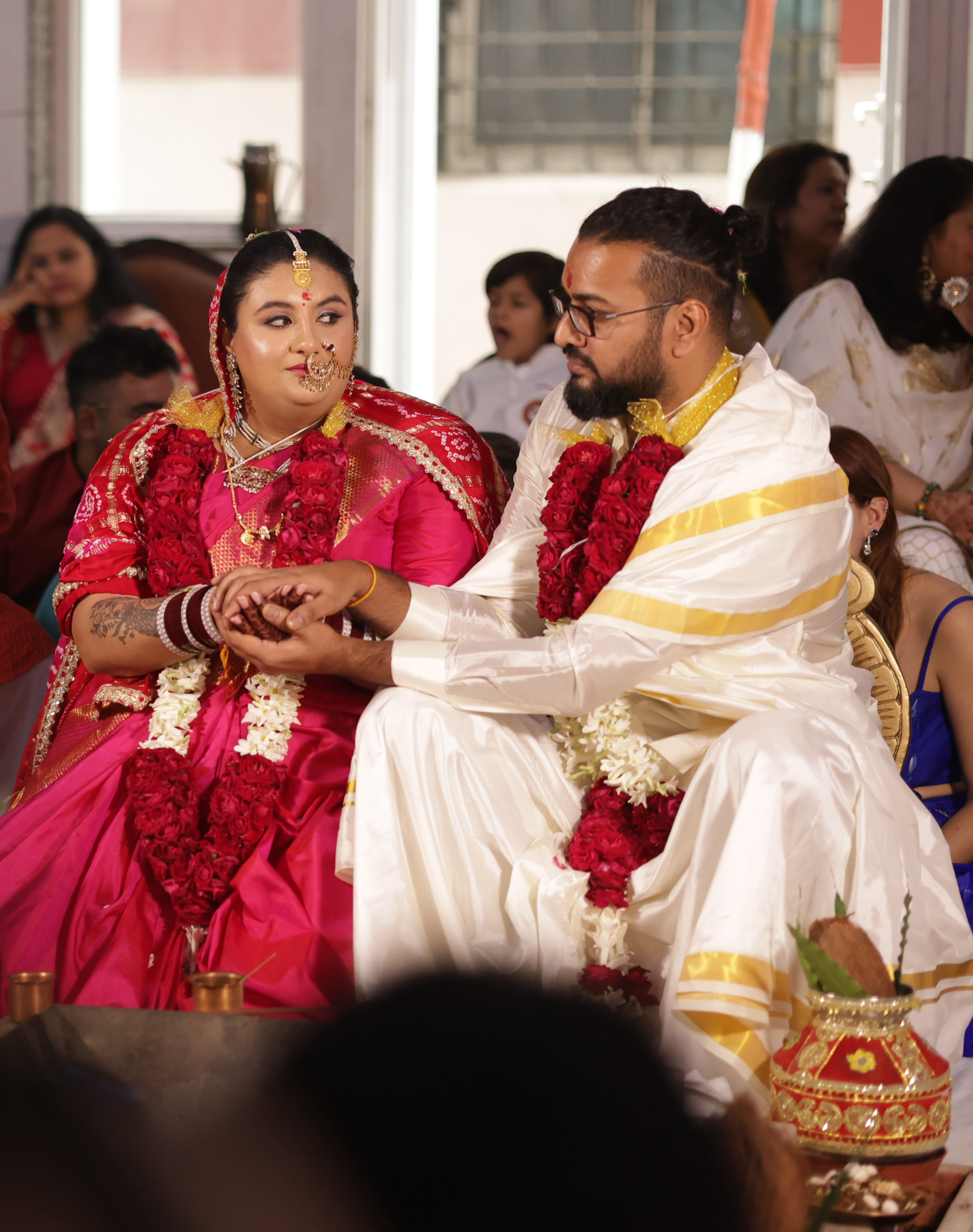 Indian wedding ceremony with a bride and groom holding hands, dressed in traditional pink and white attire, surrounded by family and friends.