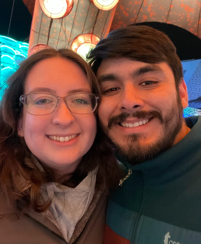 Close-up of two smiling people taking a selfie in a colorful, illuminated setting with round lighting fixtures and decorative ceiling panels.