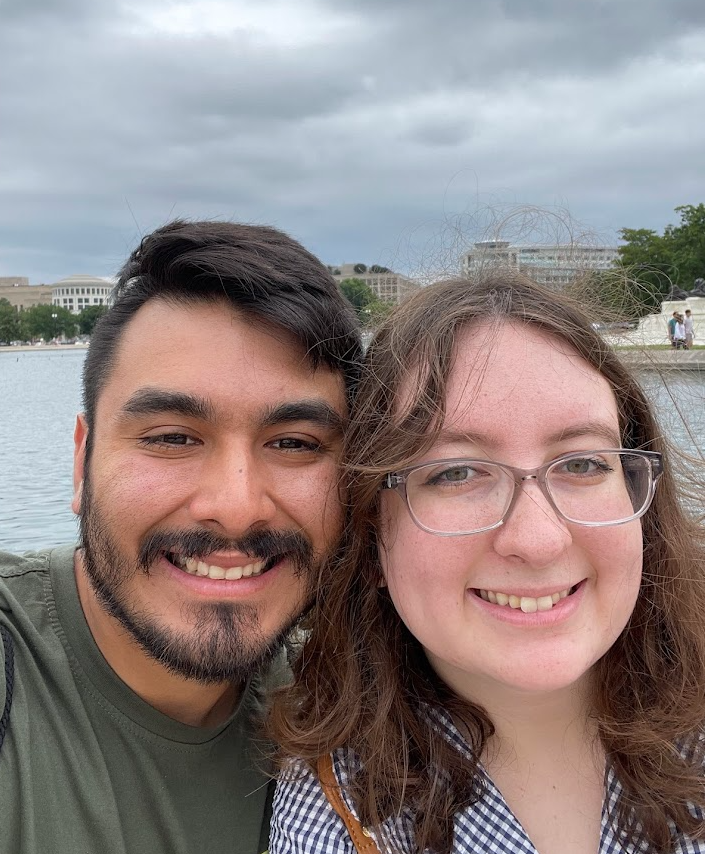 A smiling couple taking a selfie outdoors near a body of water, with cloudy skies and buildings in the background.