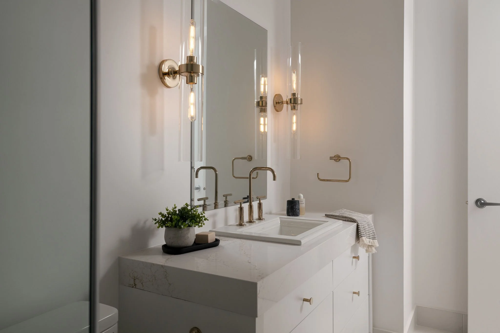 Modern white bathroom vanity with marble countertop, rectangular sink, gold fixtures, large mirror, two vertical gold light fixtures, small potted plant, and rolled towel with black and white stripes.