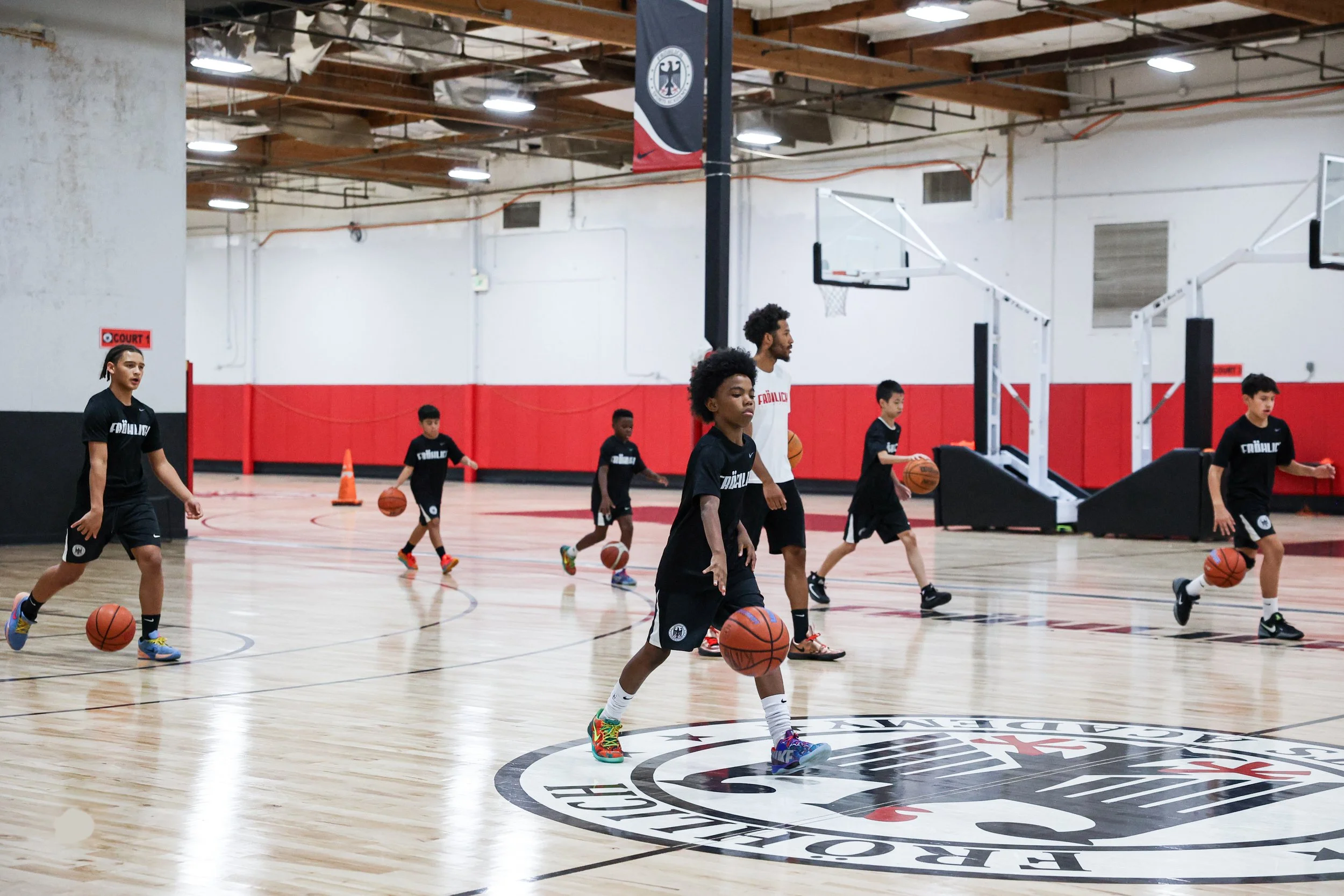 Young children practicing basketball drills in an indoor gymnasium under the supervision of an adult coach. The gym features a hardwood floor, basketball hoops, and a logo on the floor.