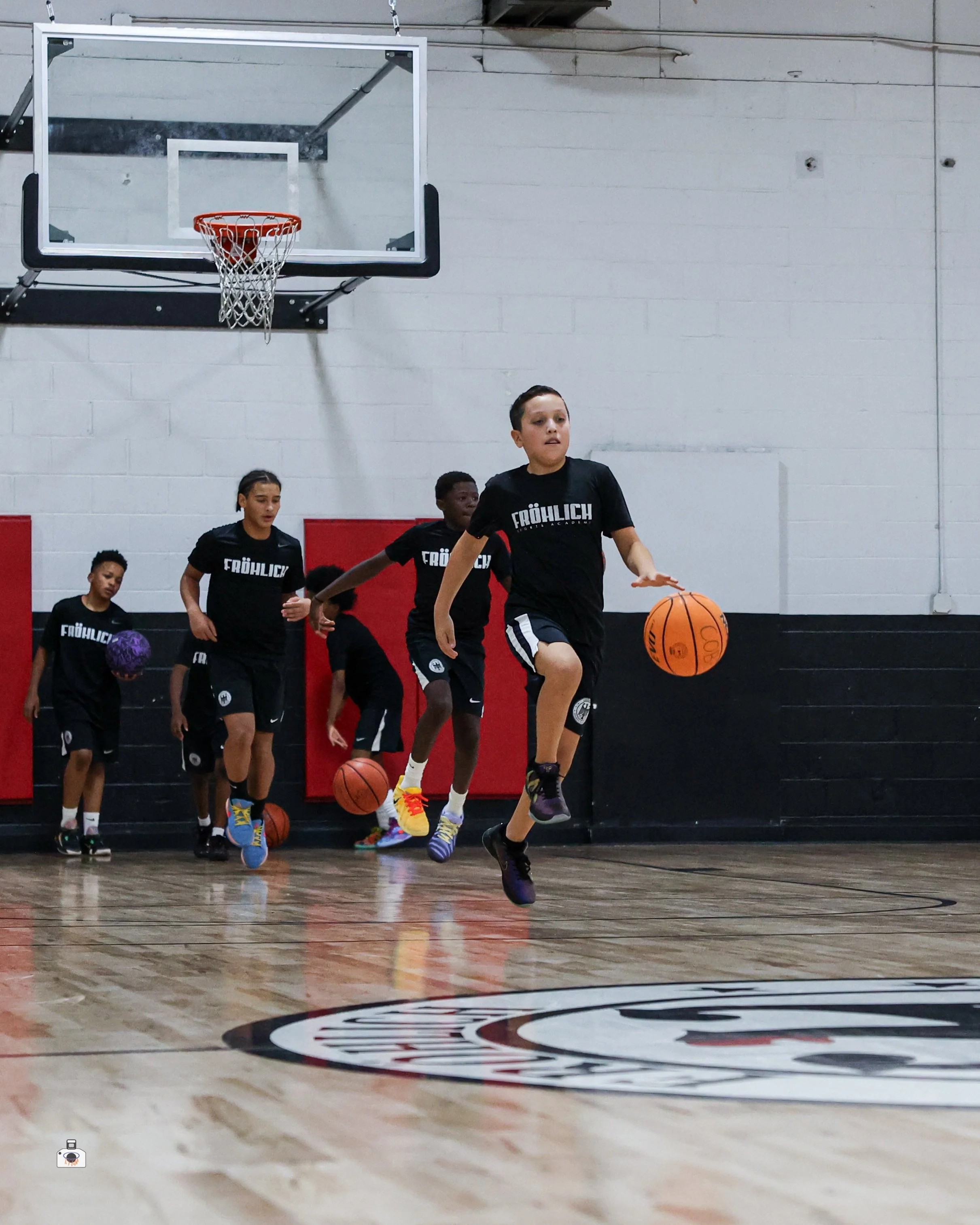 Young boys playing basketball in an indoor gym, with a basketball hoop visible on the wall. The players are wearing black shirts and are actively engaged in a game.