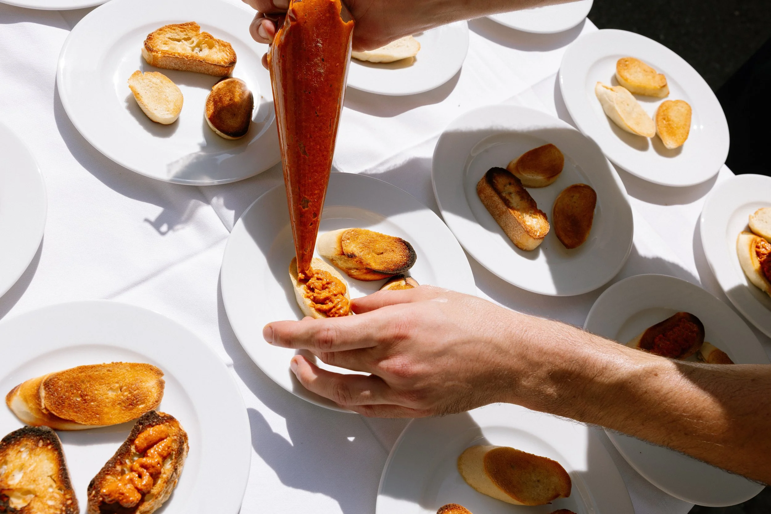 Hand, die eine Brötchenhälfte mit rotem Aufstrich bestreicht, umgeben von mehreren weißen Tellern mit verschiedenen Brötchen, auf einer weißen Tischdecke bei Tageslicht.