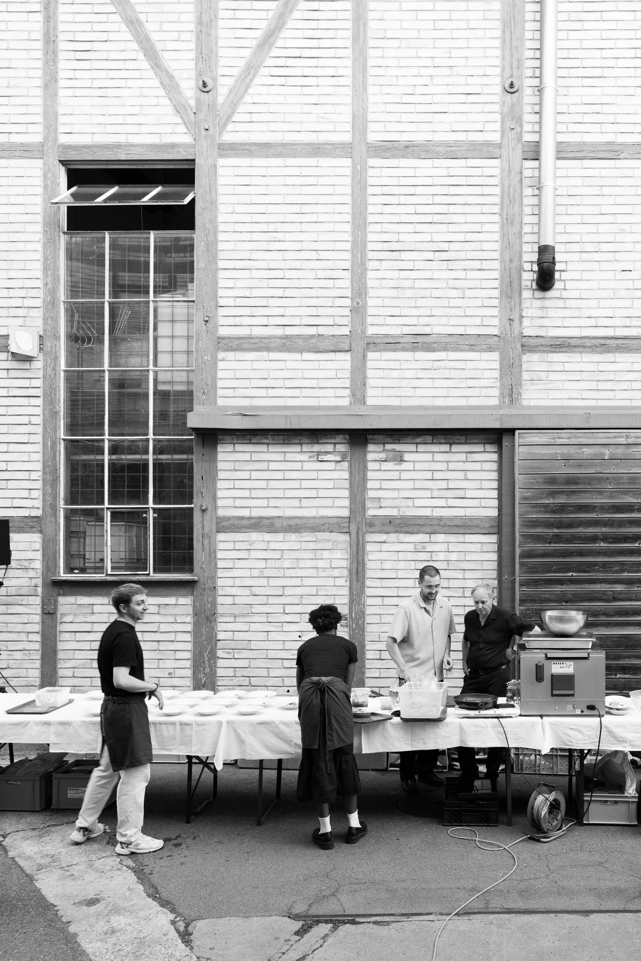 Menschen beim Grillen und Zubereiten von Essen unter einer offenen Holz- und Ziegelfassade