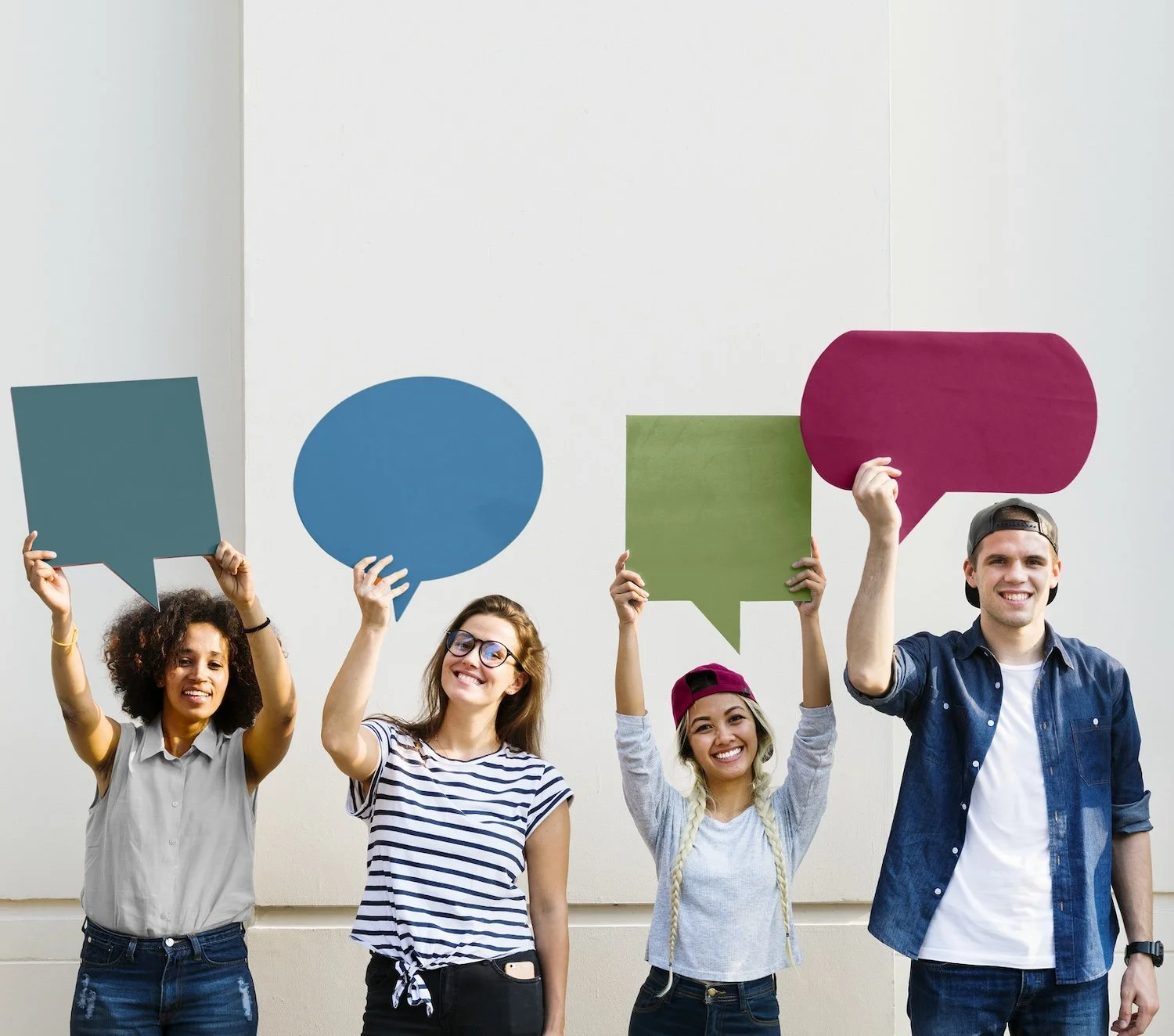 Group of four young people standing against a plain wall, holding colorful speech bubble signs and smiling.