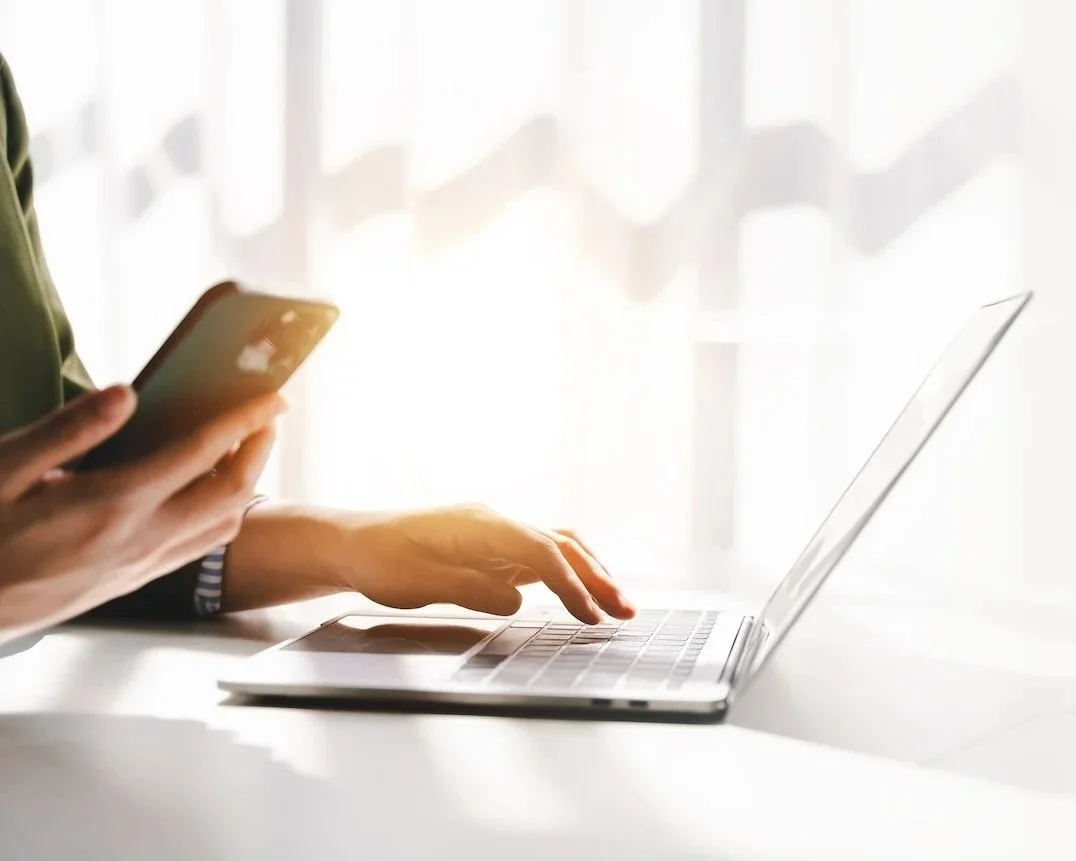 woman holding phone and typing on laptop