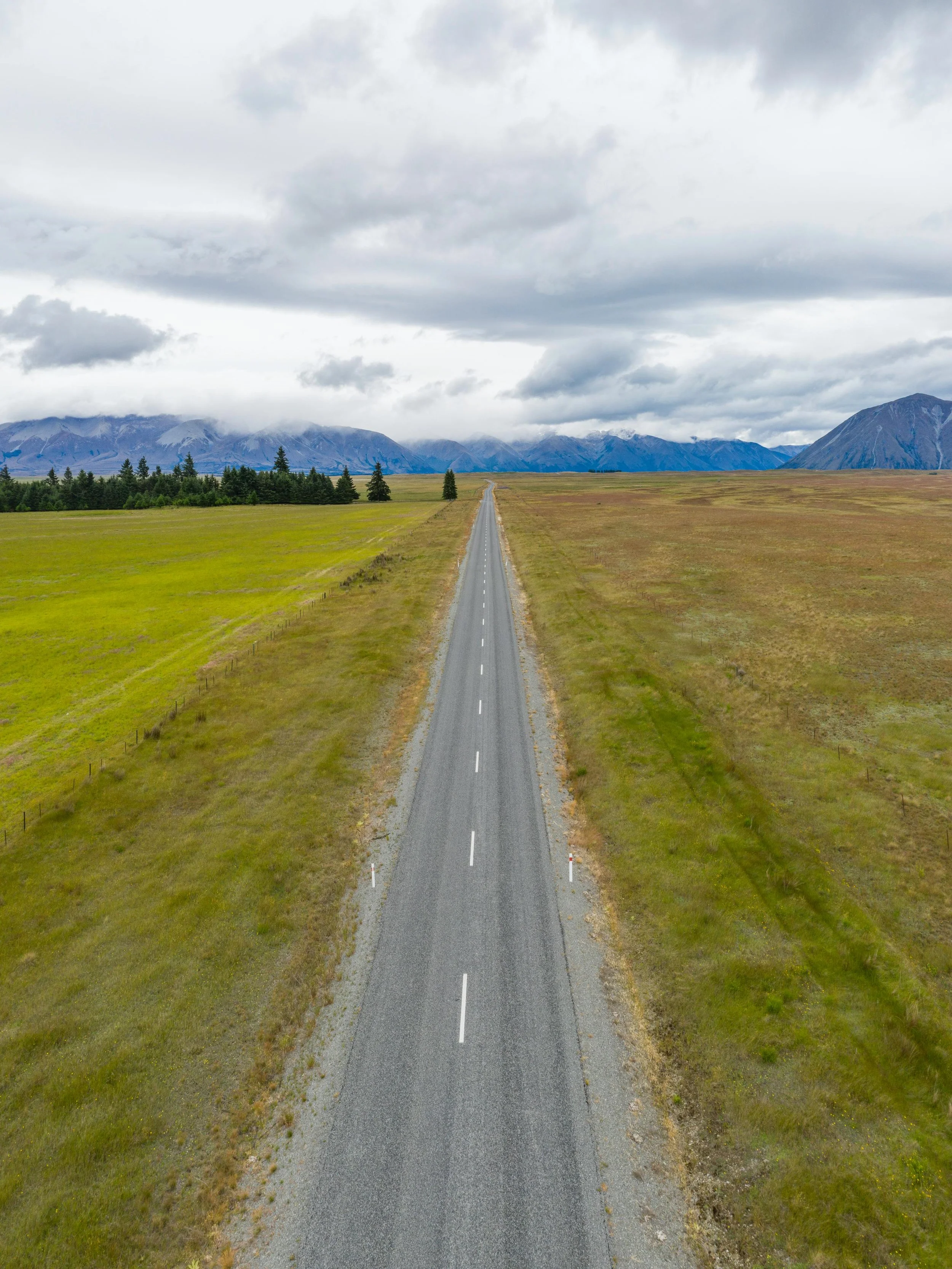 A straight road running through open grassy fields under cloudy sky, with distant mountains in the background.