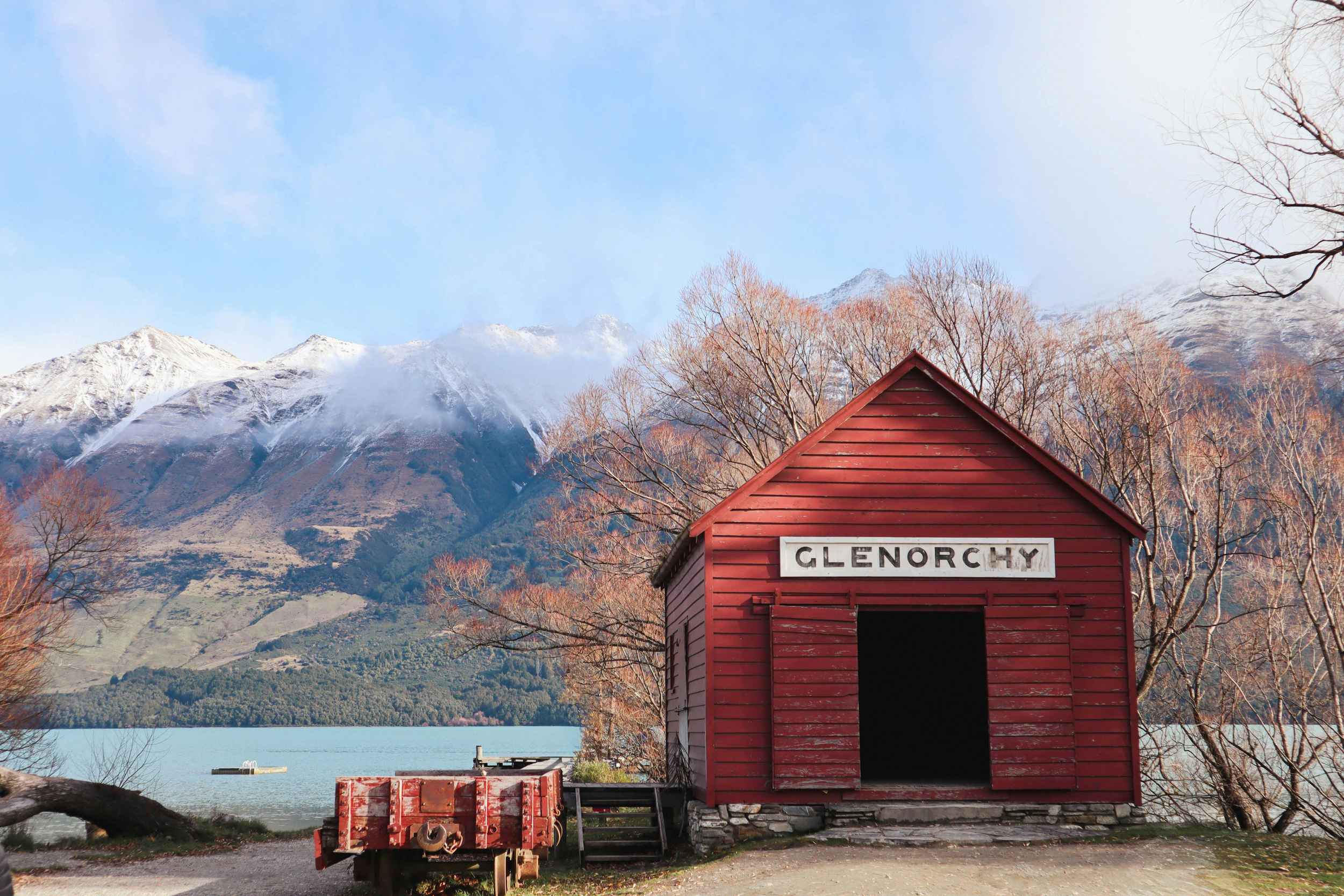 A red wooden building labeled 'Glenorchy' with a snow-capped mountain in the background, trees with bare branches, and a body of water with a barrel and a platform in the foreground.