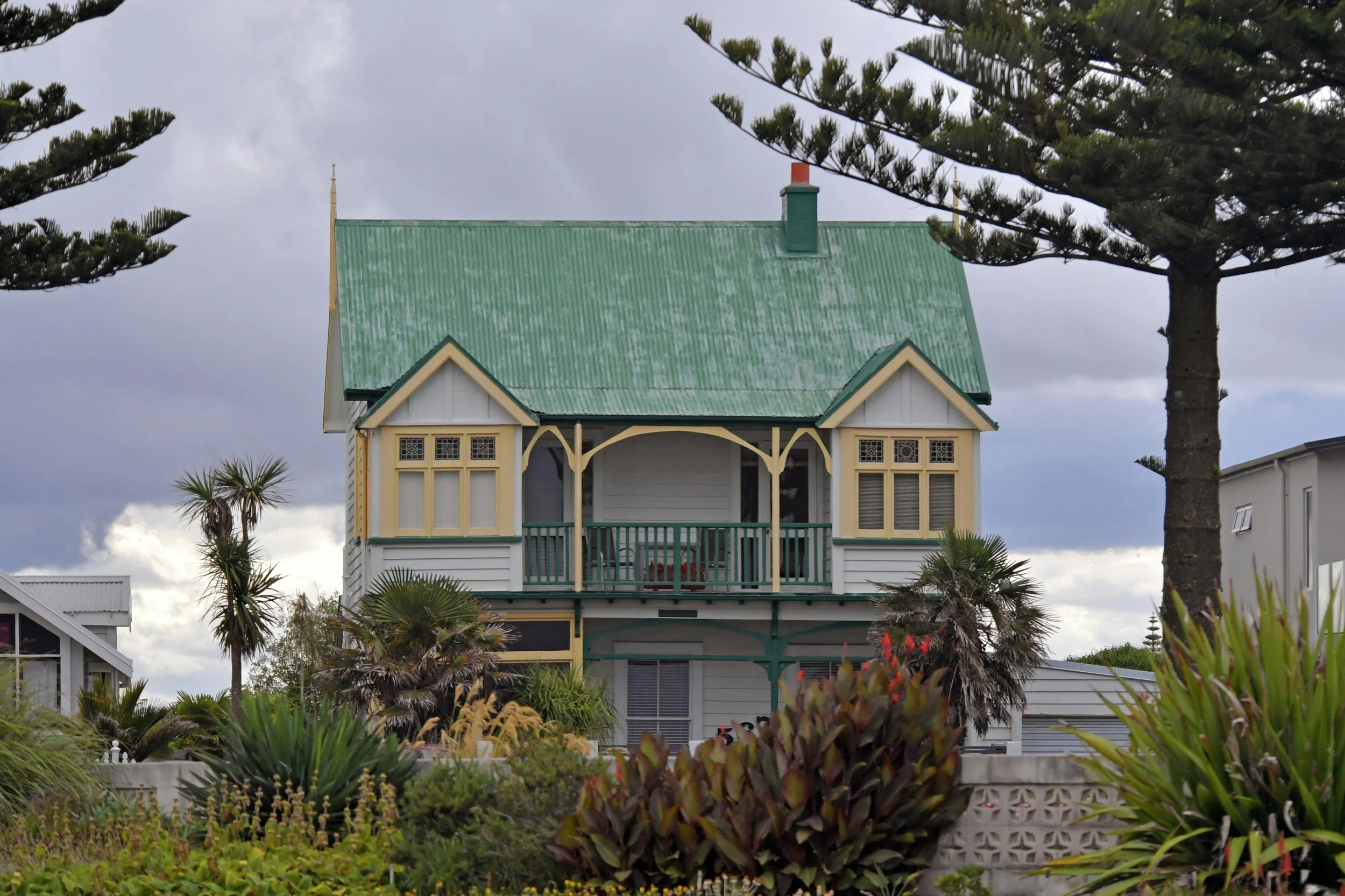 A two-story house with a green sloped roof, cream trim, and a front porch. The house is surrounded by lush green plants and trees, with a cloudy sky overhead.