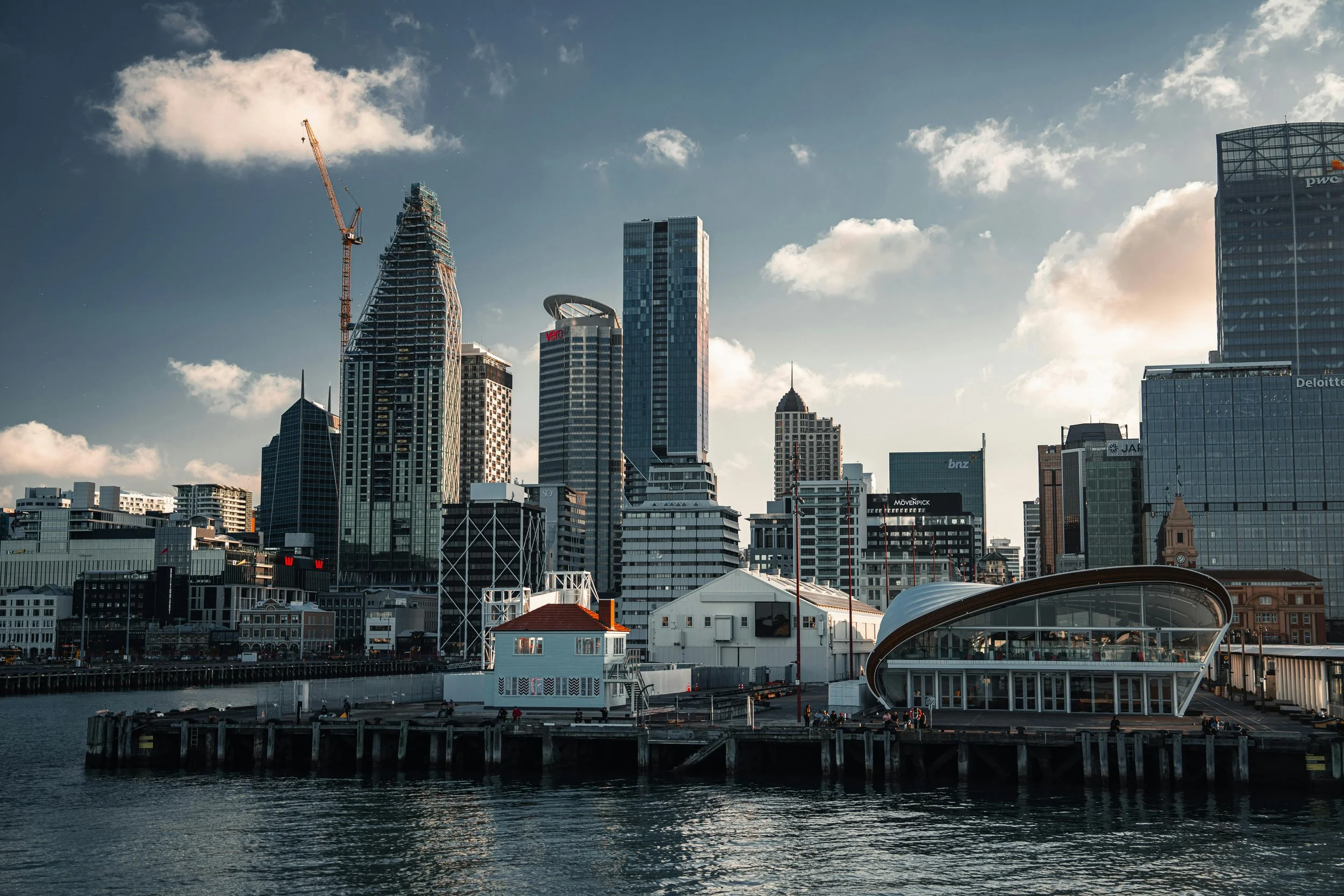 City skyline with modern skyscrapers under a partly cloudy sky, viewed from the water's edge with a wooden pier and a contemporary building extending over the water.