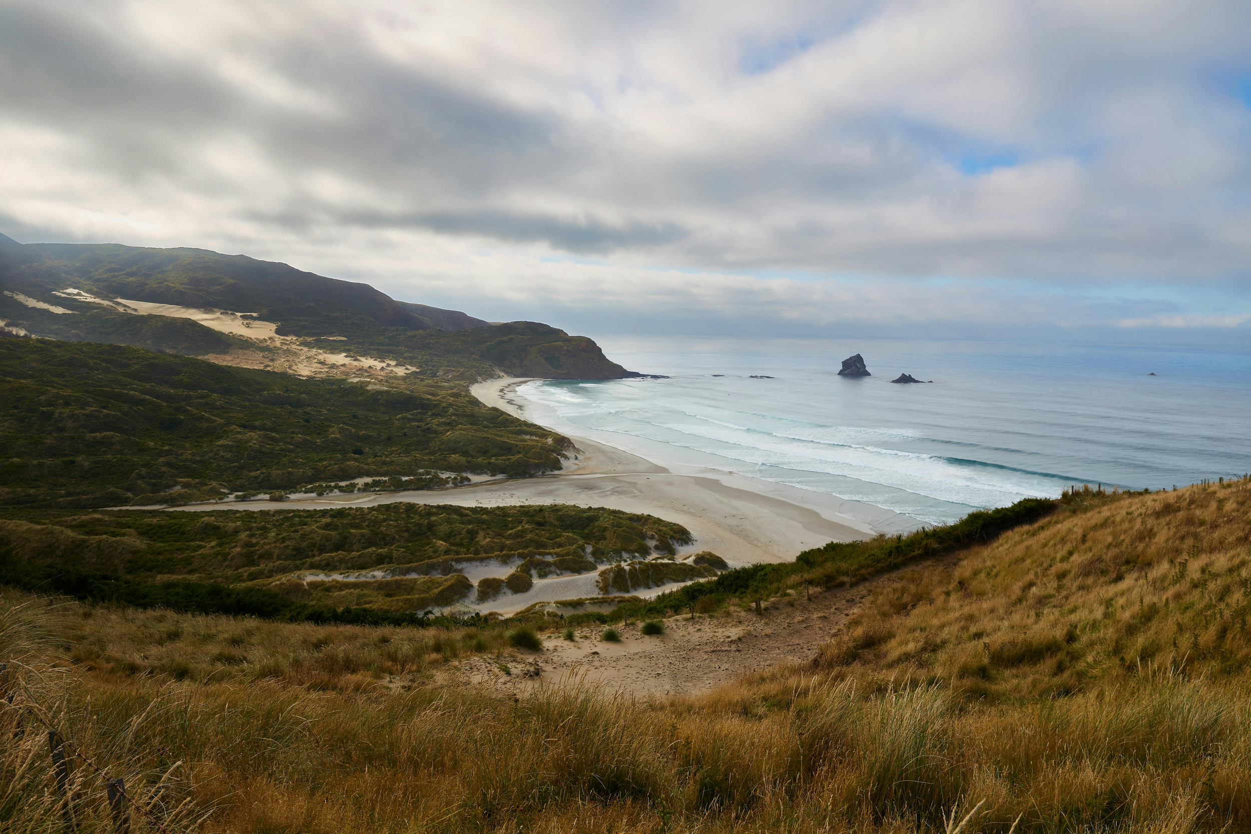Scenic view of a sandy beach bordered by green hills and ocean waves, with a rocky formation in the distance under cloudy sky.