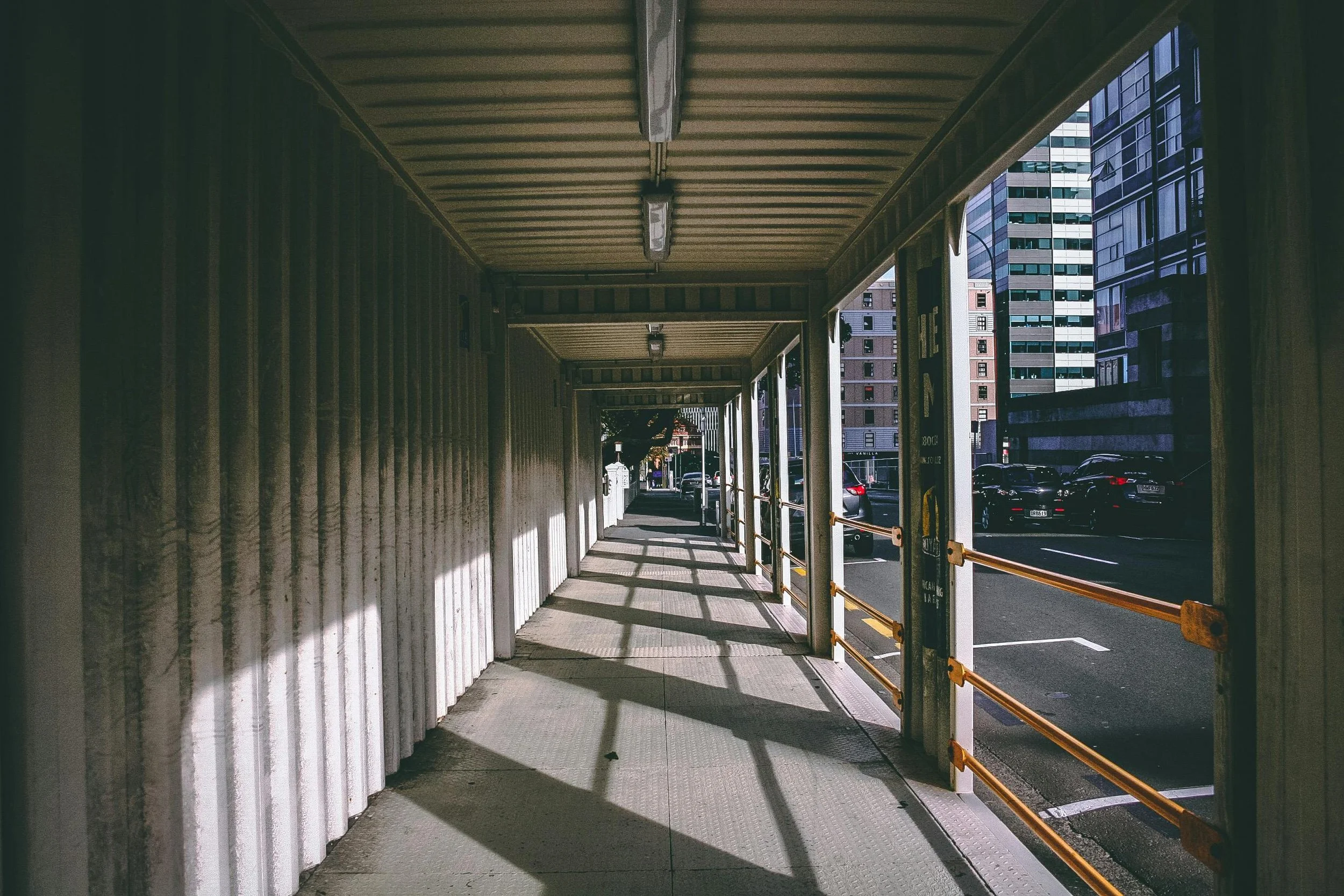 Urban sidewalk with construction barriers, shadows cast by the overhead covering, and tall buildings in the background.