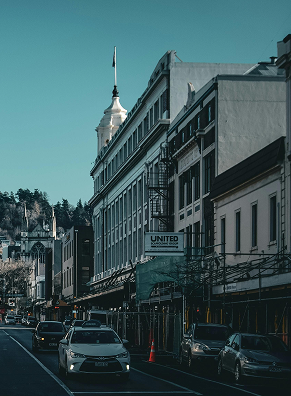 Street view of historic buildings with a clock tower in the background, cars parked along the road, and a clear blue sky.