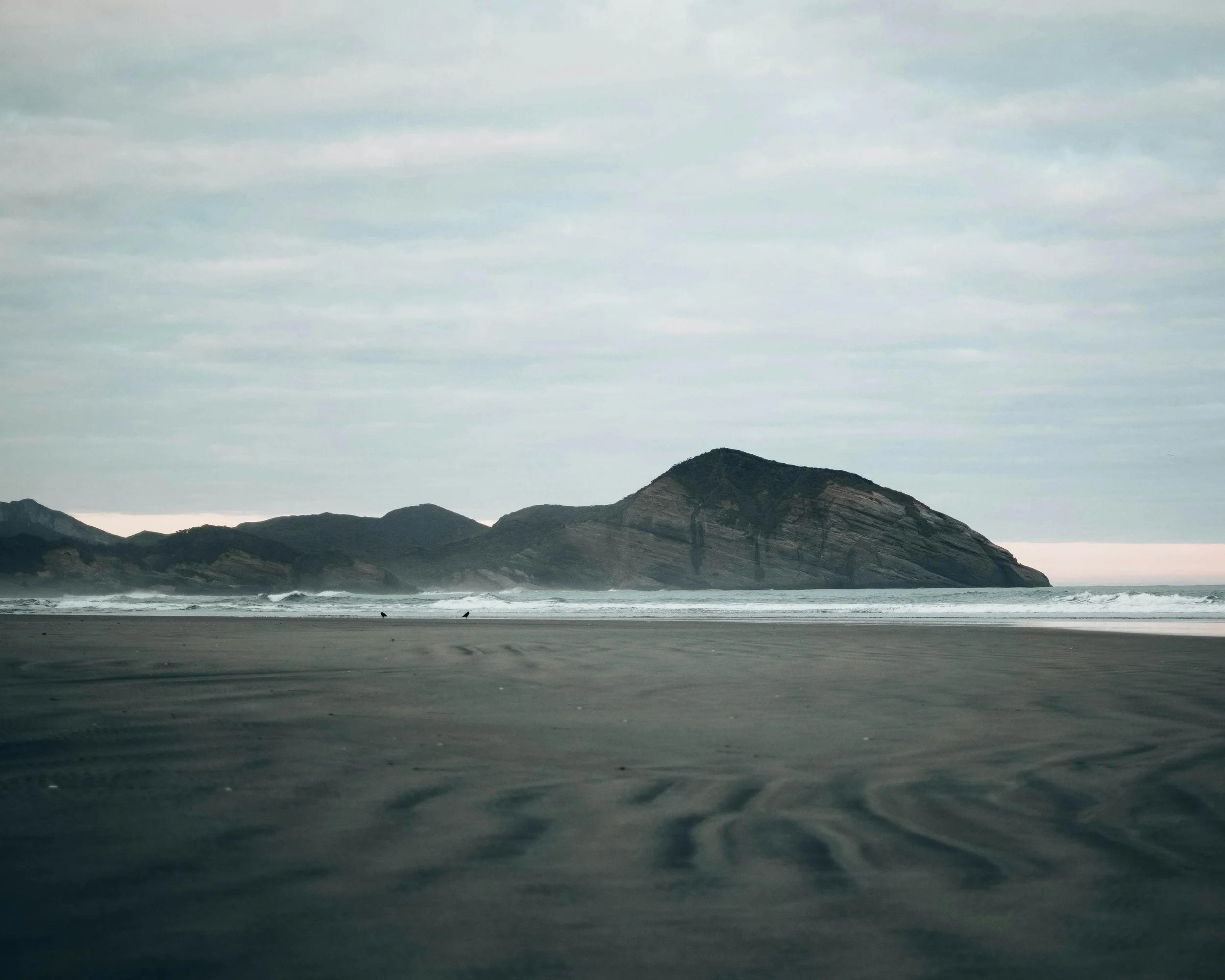 A beach with dark sand, ocean waves, and a mountain in the distance under a cloudy sky.