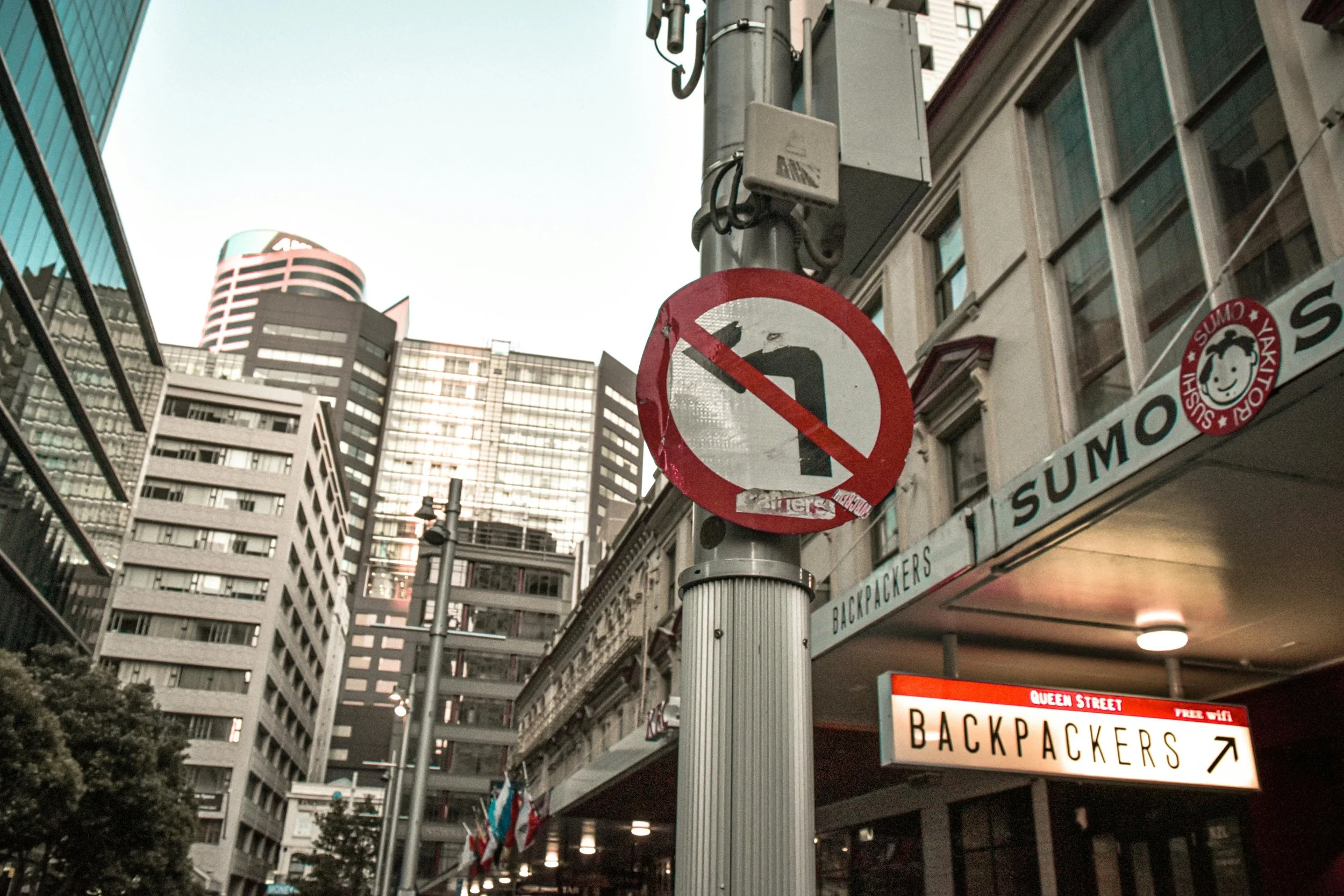 No left turns sign on a city street pole with tall buildings and a sign pointing to backpackers on Queen Street.