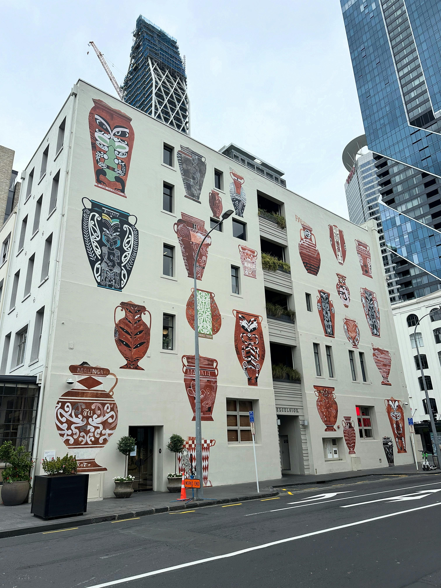 A white multi-story building decorated with colorful indigenous art murals of various vases and pottery on its exterior wall, located in an urban area with high-rise buildings and a street with a traffic cone in the foreground.