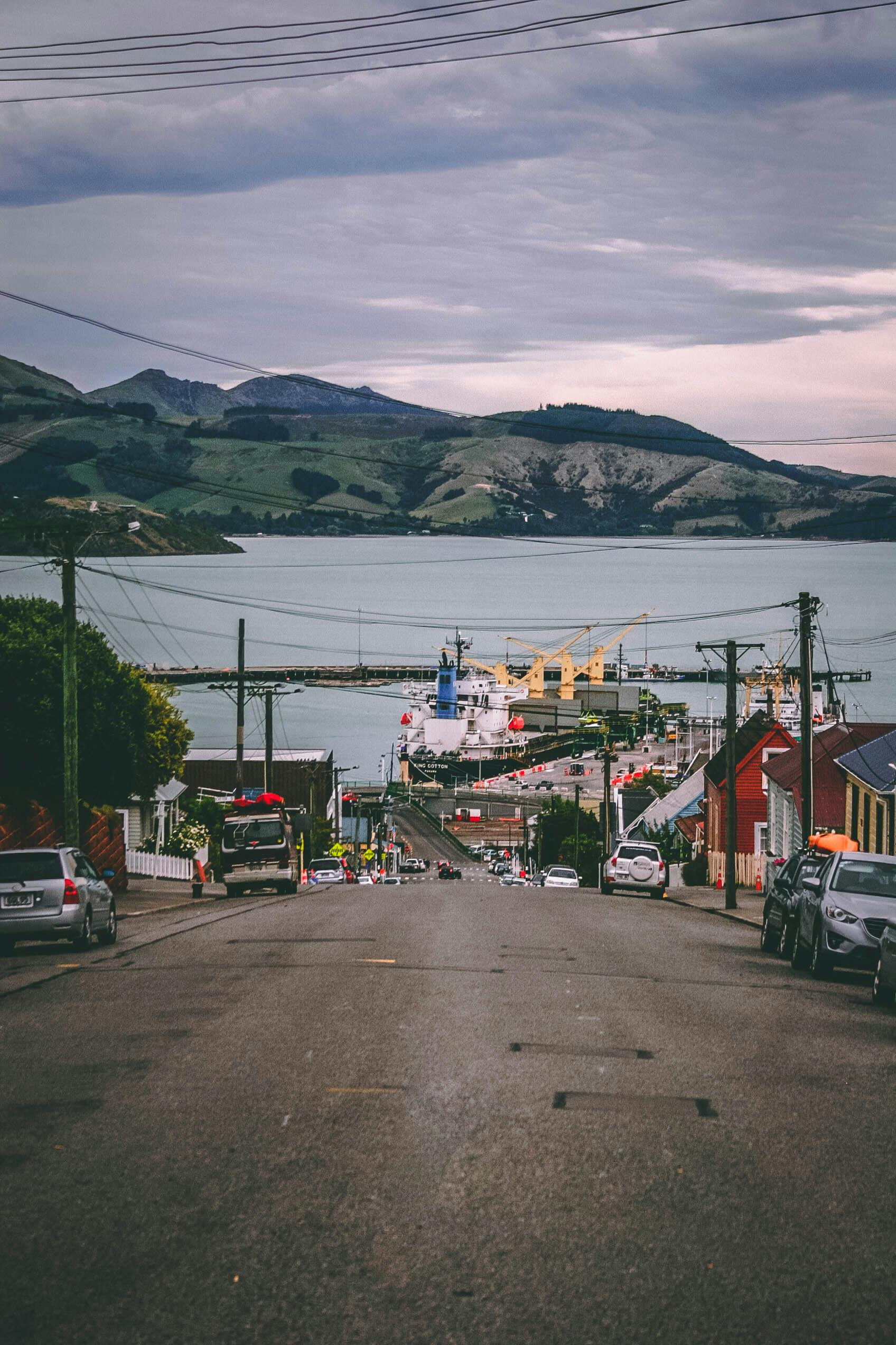 A hilly coastal town with buildings lining a street leading to a harbor with ships and cranes, overlooking a body of water and mountains in the background.