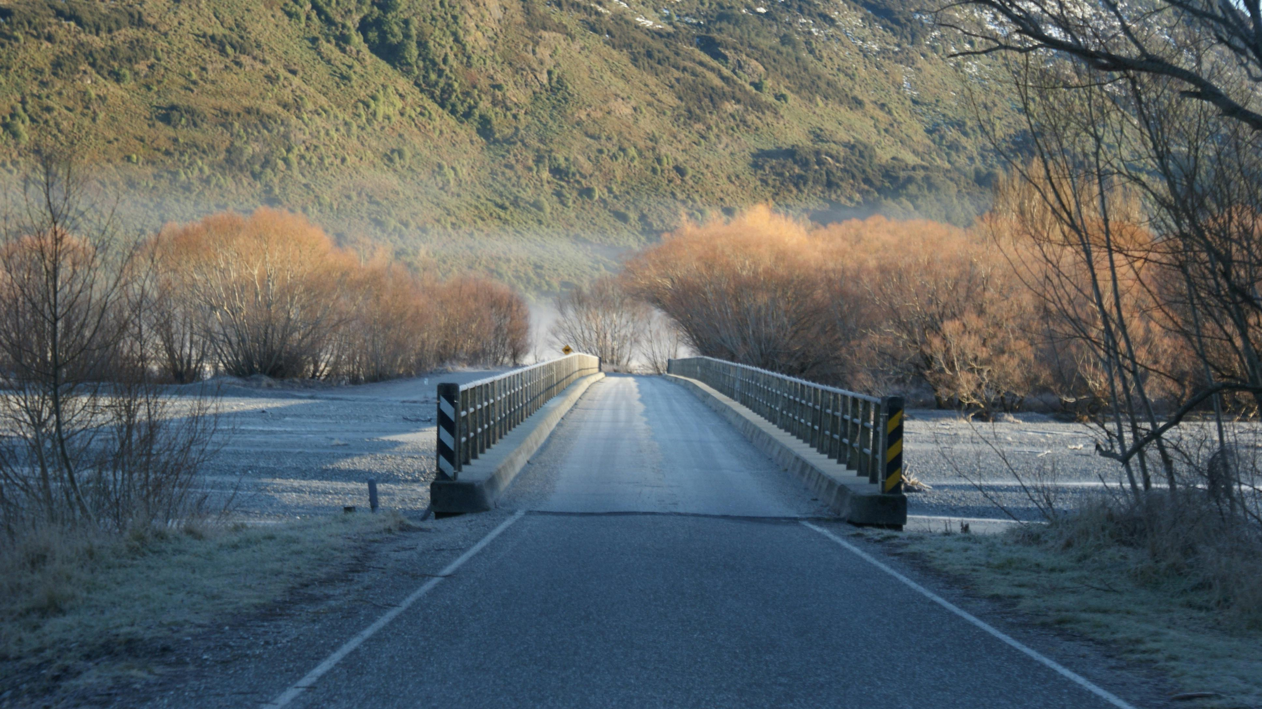 A narrow road leading to a small bridge over a frozen river, with leafless trees and hilly terrain in the background, during winter.