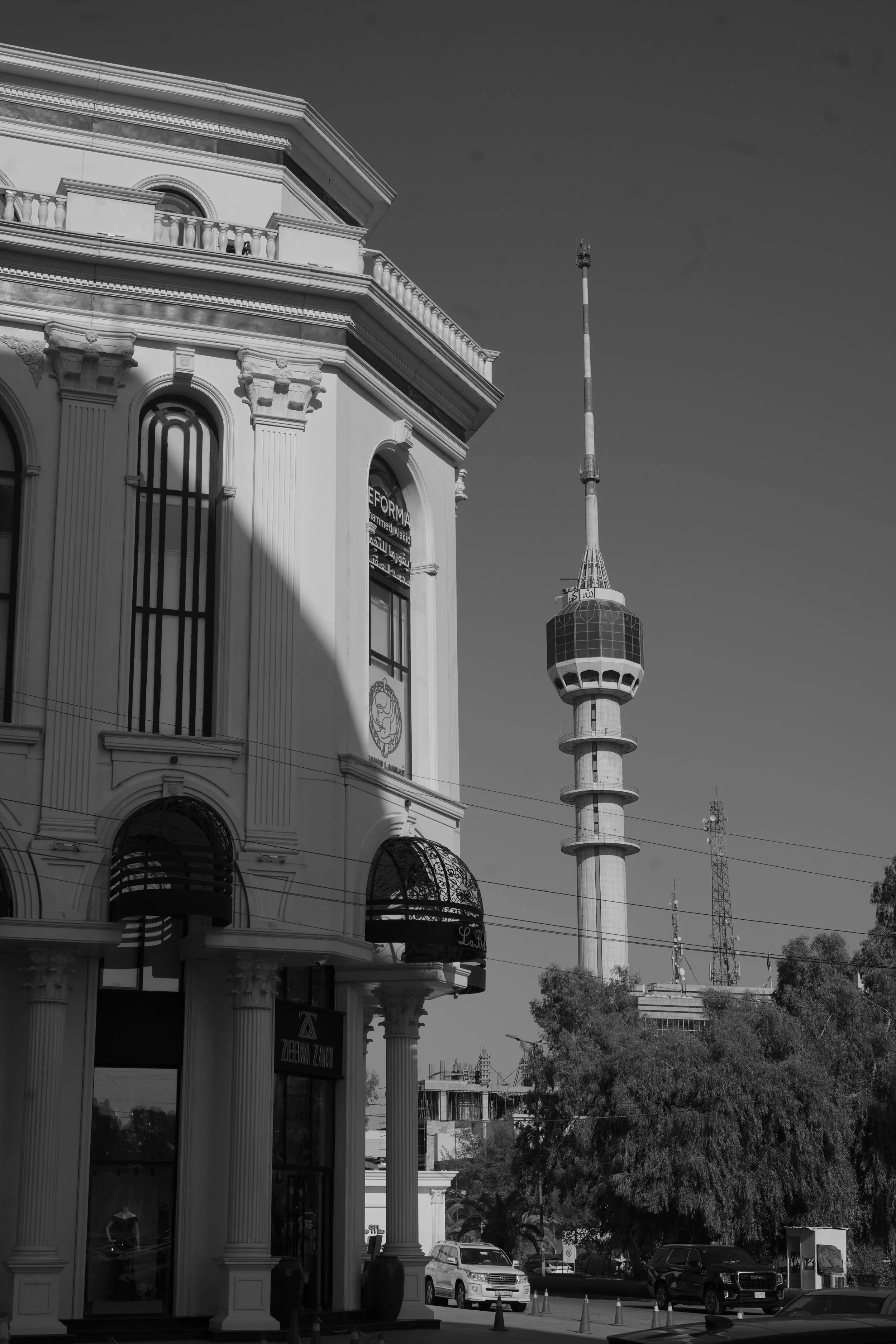 Cityscape featuring a large classical style building with columns and arched windows in the foreground and a tall telecommunications tower in the background under a clear sky.