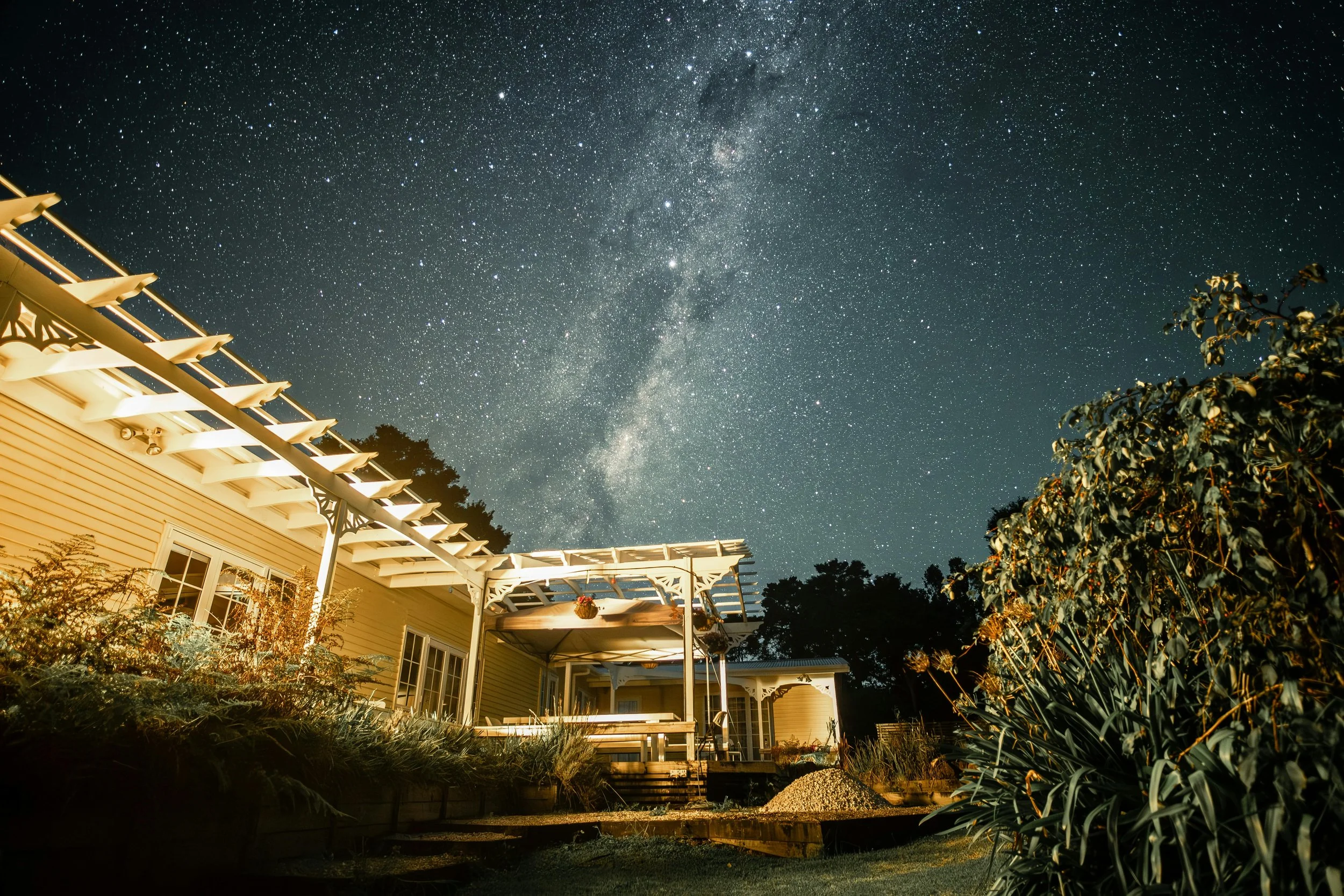 Night sky filled with stars and the Milky Way above a house with a lit patio and garden plants.