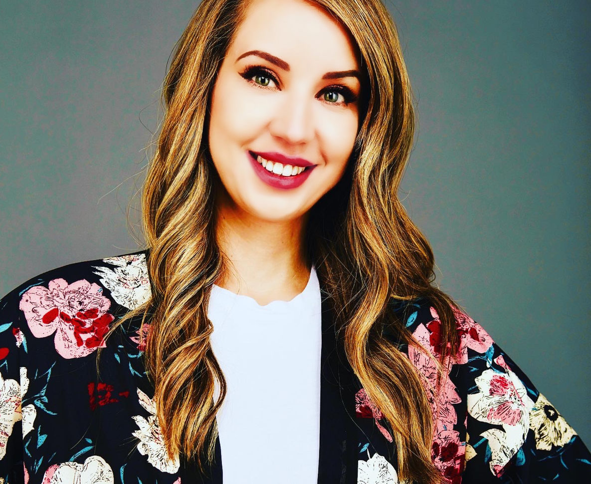 A smiling woman with long wavy hair, wearing a floral patterned black jacket over a white top, with a neutral gray background.
