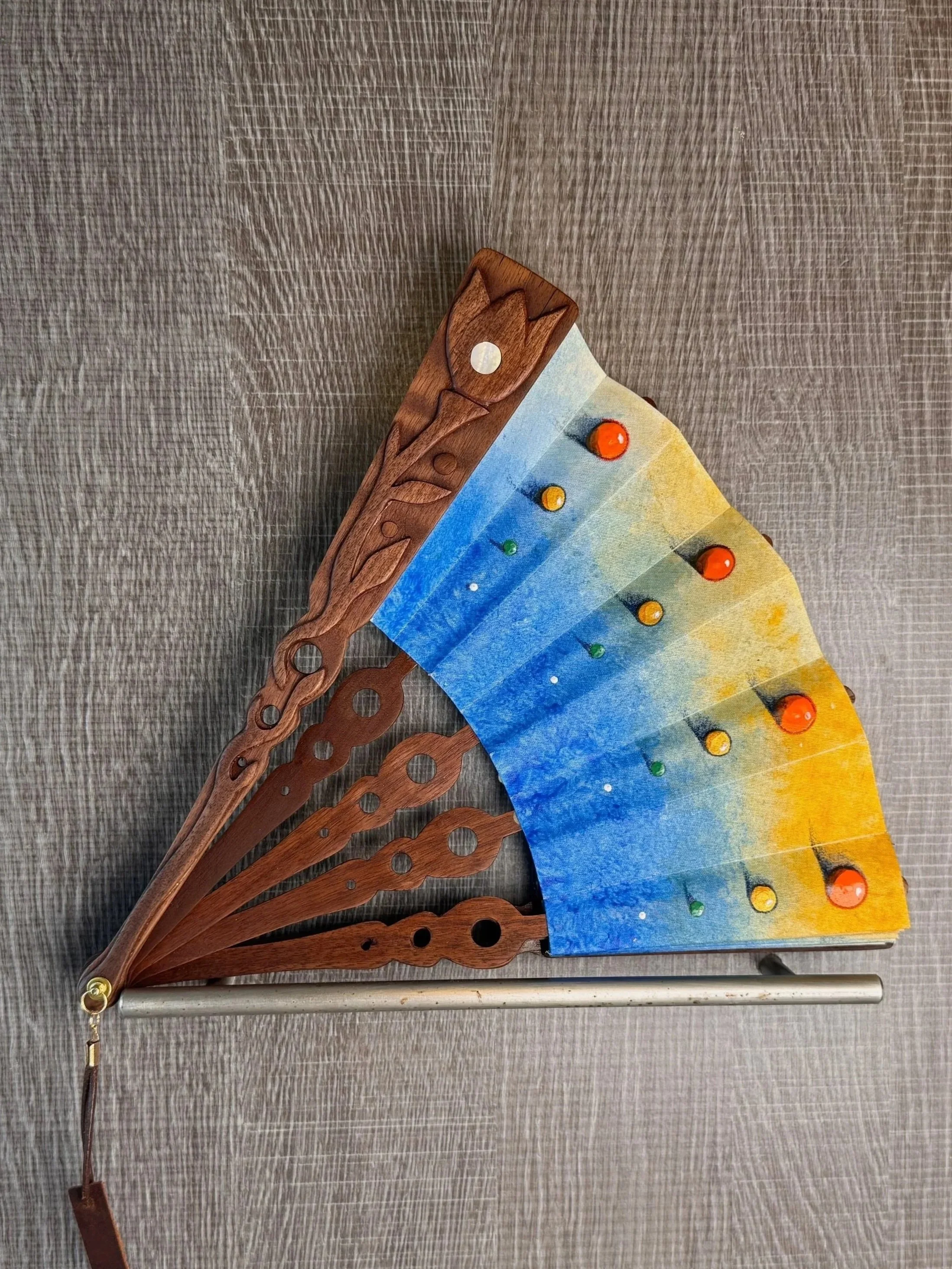Colorful hand fan with wooden ribs and painted fabric, featuring a blue to yellow gradient, and red, yellow, and green decorative elements.