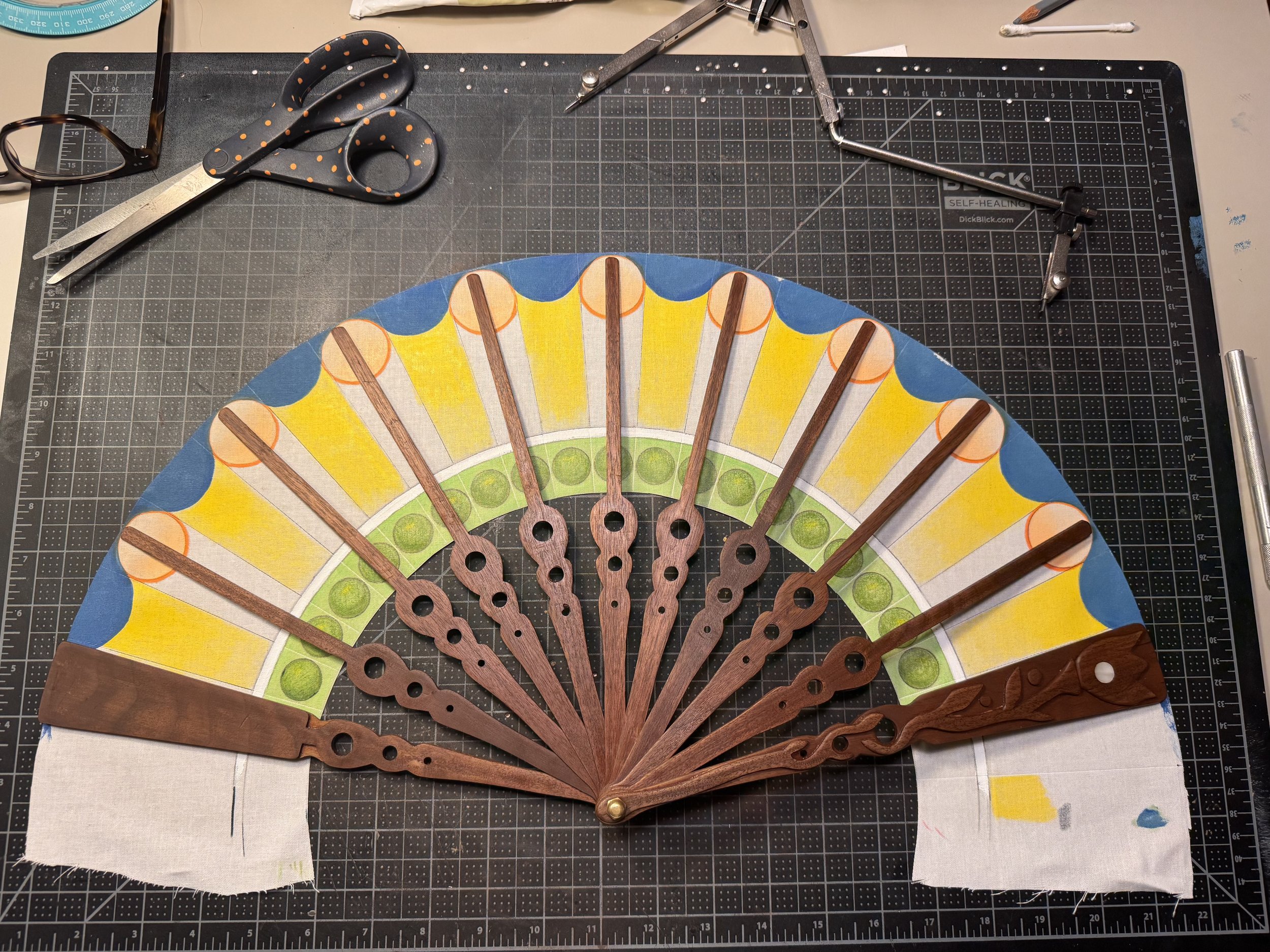 A handcrafted fabric fan with wooden slats, painted with colorful designs, is laid out on a work surface. The fan is partially assembled with the slats attached at the base. Around the fan are drafting tools, scissors, and a cutting mat.