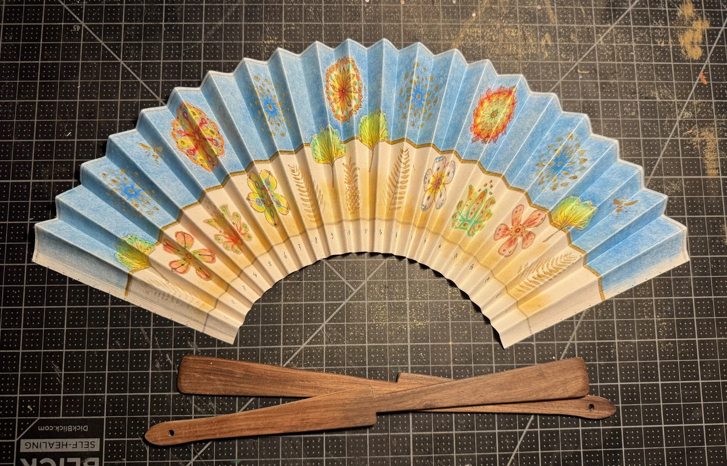 Decorative hand fan with blue and beige panels featuring butterfly and floral designs, placed on a crafting grid surface with wooden sticks below.
