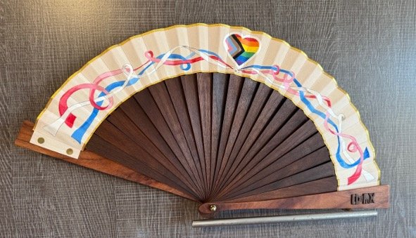 Decorative hand fan with a rainbow-colored heart and multi color ribbons design on a white background, lying open on a dark wooden surface.