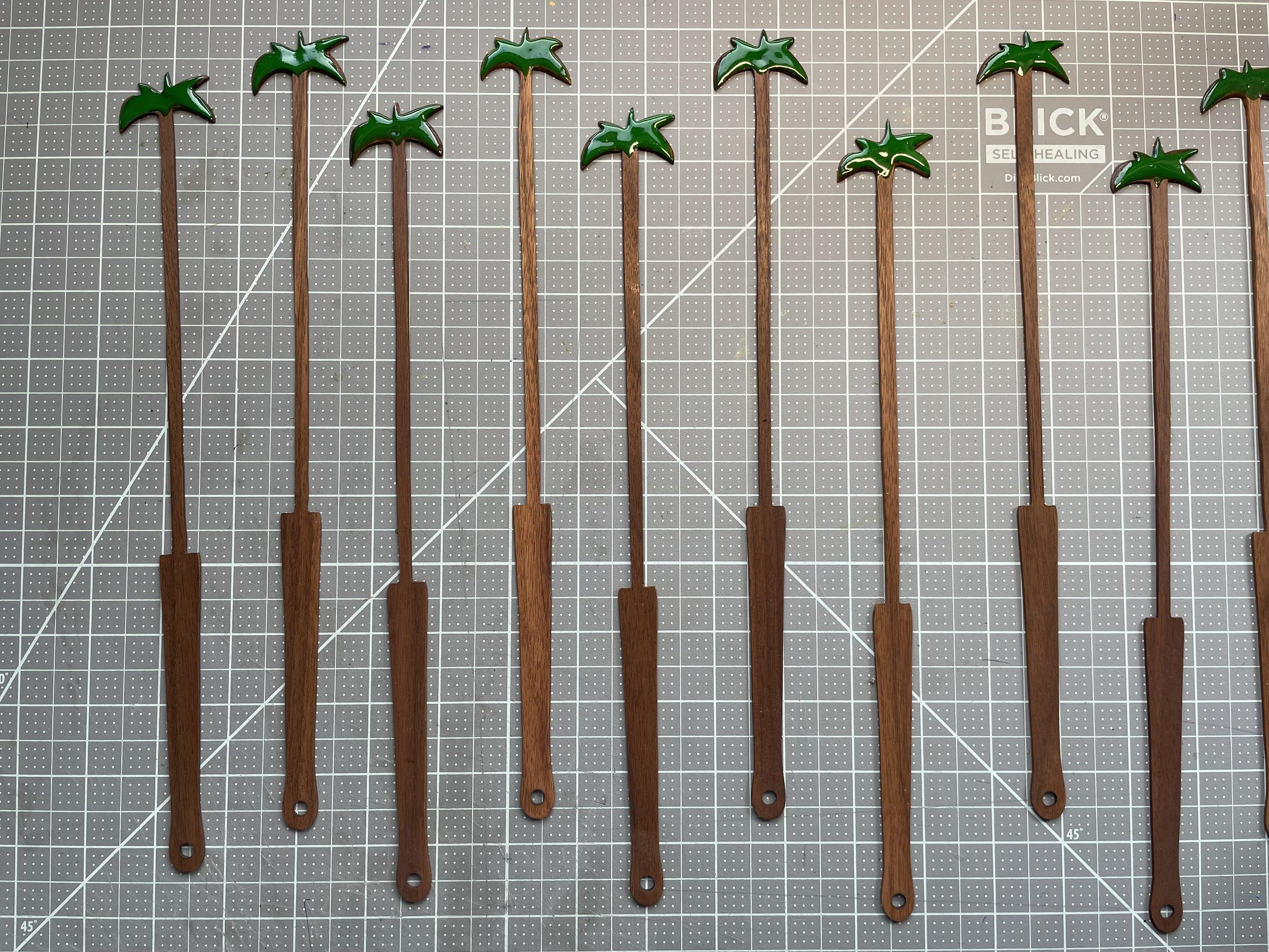 Ten carved fan staves with green palm tree decorations attached to the top of each, laid out on a gray cutting mat.